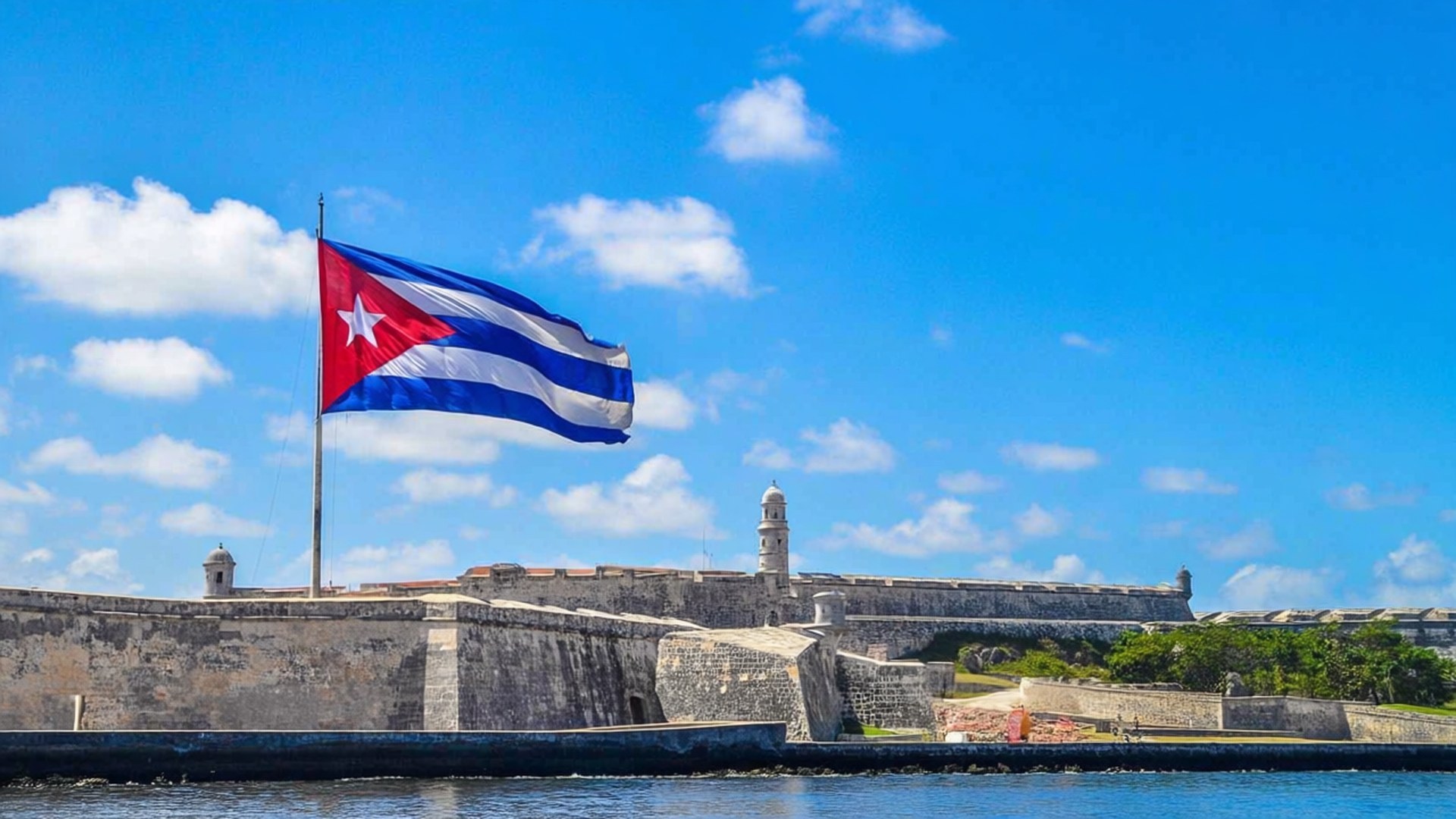 Flag at El Morro Castle 壁紙 — 古巴國旗在歷史悠久的石頭堡壘頂部自豪地飄揚，俯瞰著平靜的藍色水域，天空明亮，雲朵散落。