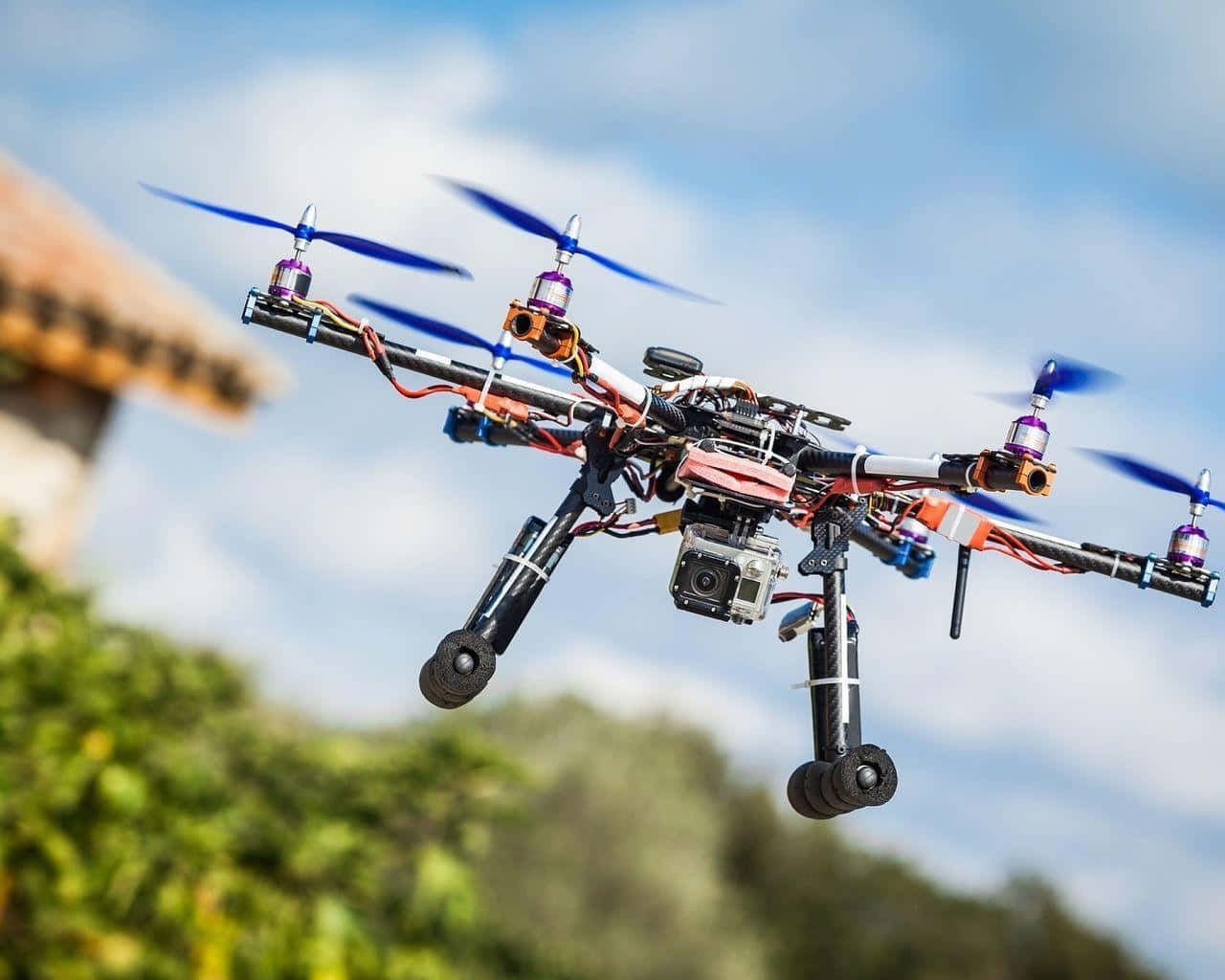 Drone Camera 桌布 壁紙 — 一架自製的四軸無人機，帶有外露的接線和相機，在明亮的藍天下懸停，下方是模糊的綠色植物。