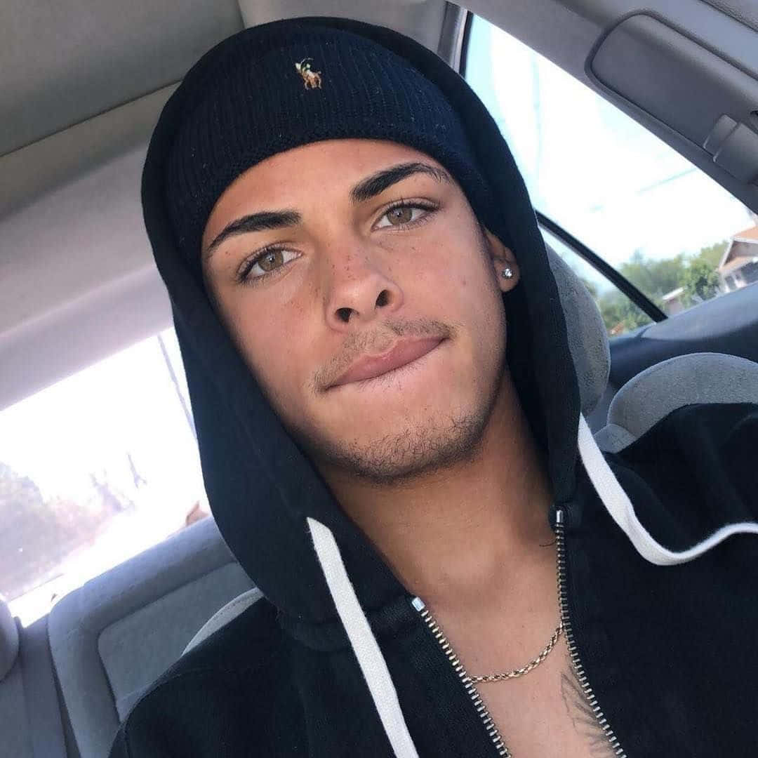  壁紙 — 一個戴黑色毛帽和連帽上衣的年輕男子坐在車內，直視鏡頭，露出微妙微笑。