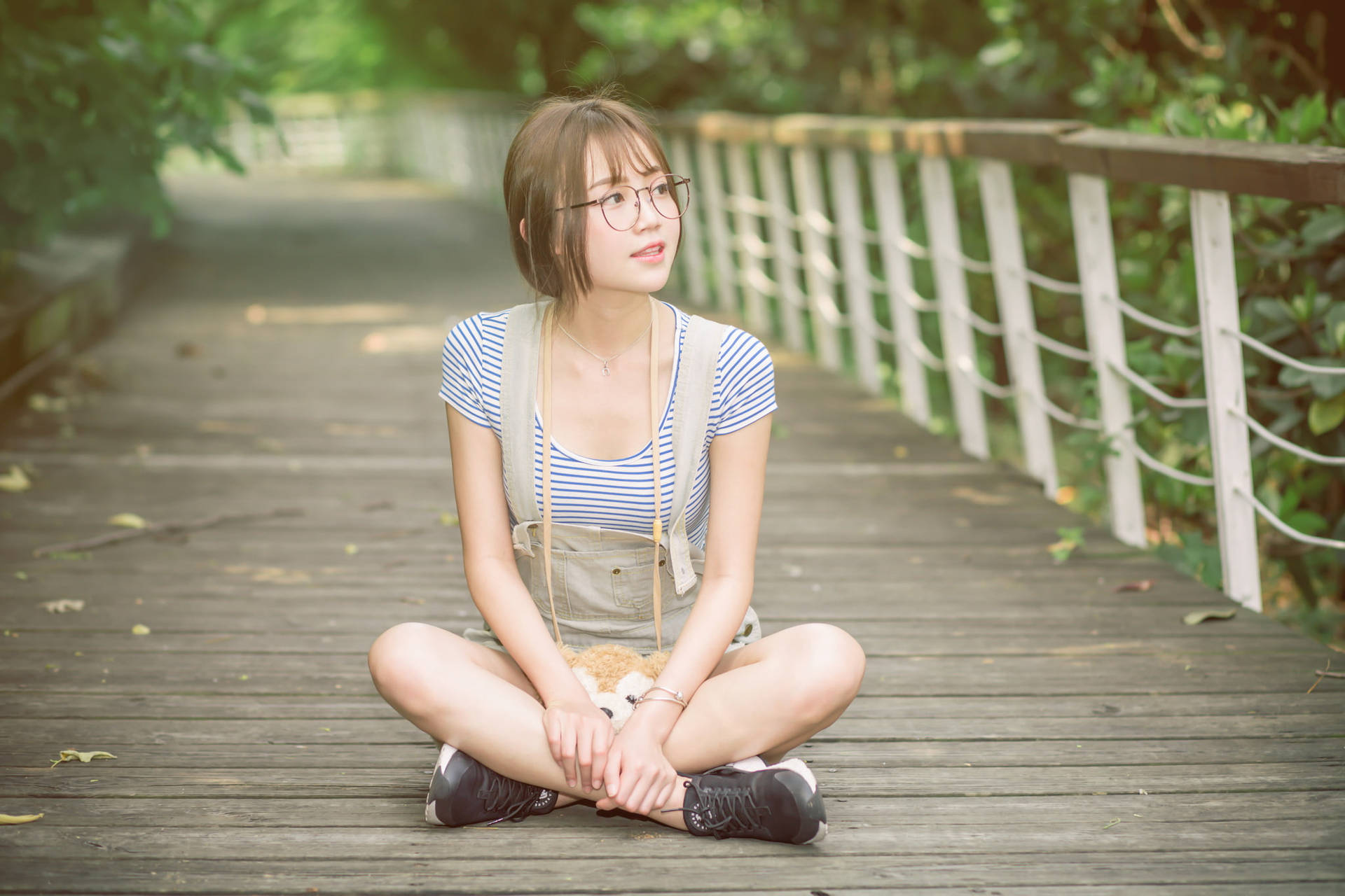 戴眼鏡的年輕女性盤腿坐在公園的木製步道上。 — 日式自然壁紙 壁紙
