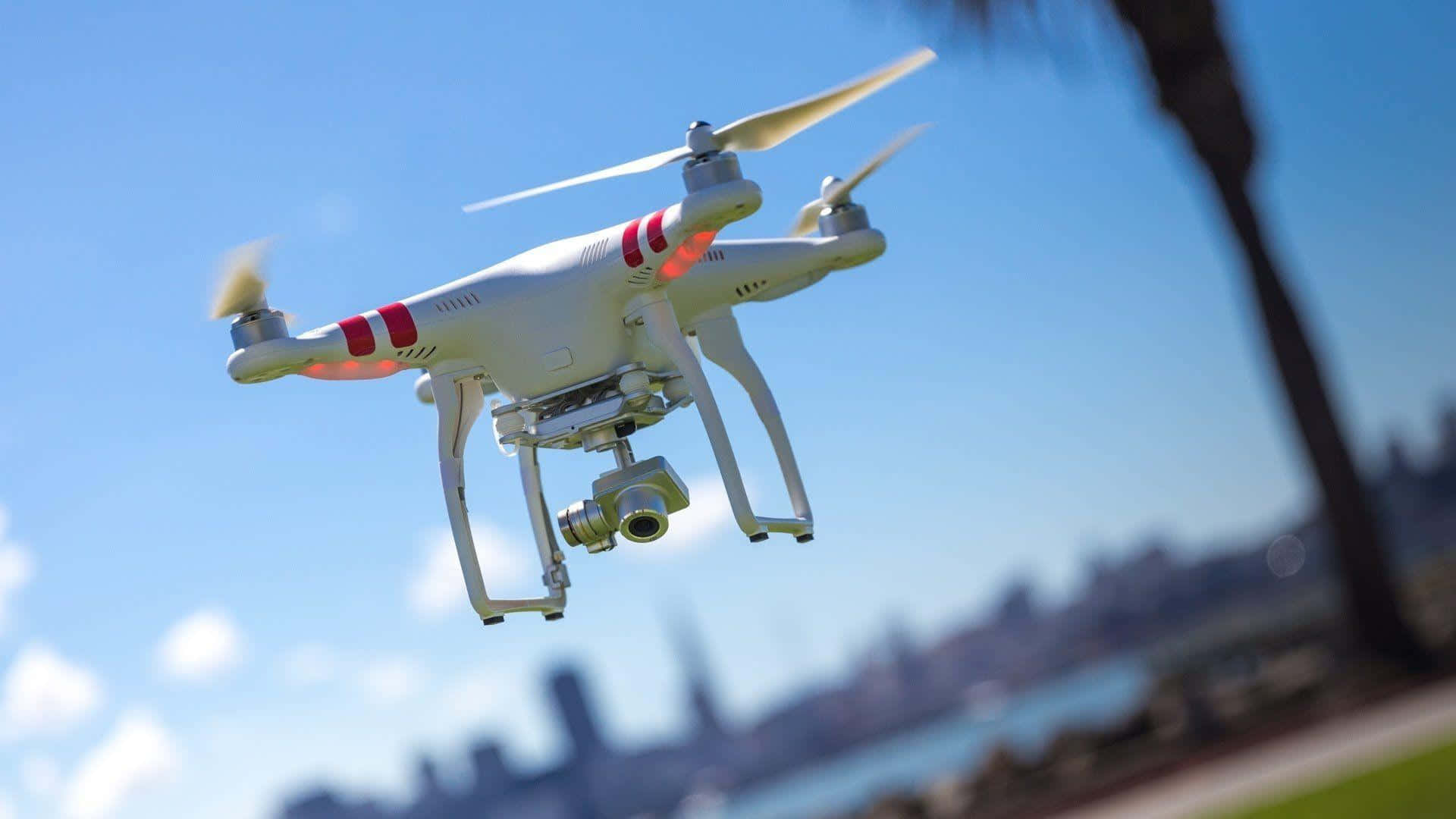 Drone Camera 桌布 壁紙 — 一架帶有紅色條紋的白色無人機在清澈的藍天中盤旋，背景是模糊的城市景觀和棕櫚樹。