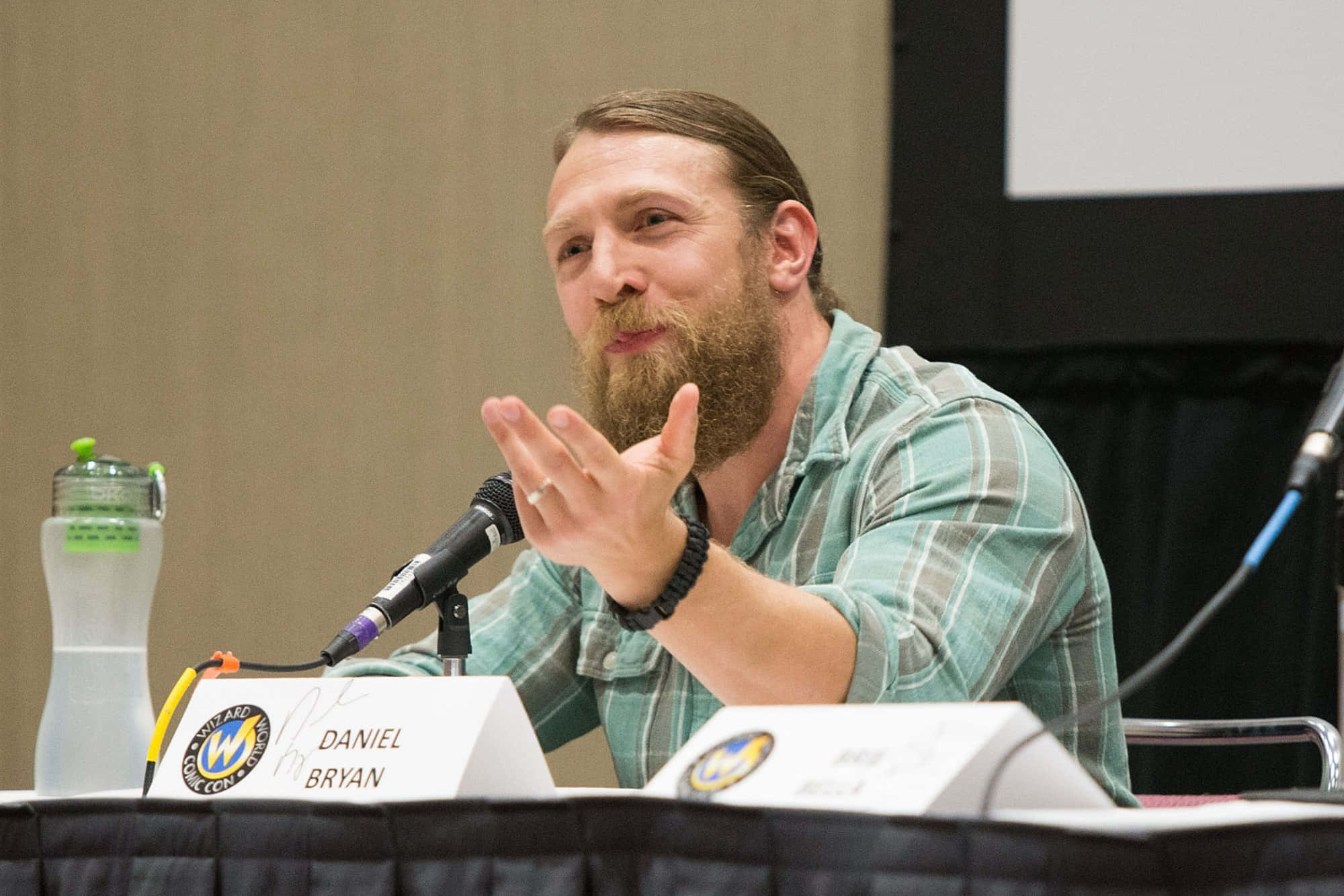 摔角 桌布 壁紙 — Daniel Bryan 在Wizard Con座談會上生動地比劃手勢，桌上放著麥克風和水瓶。