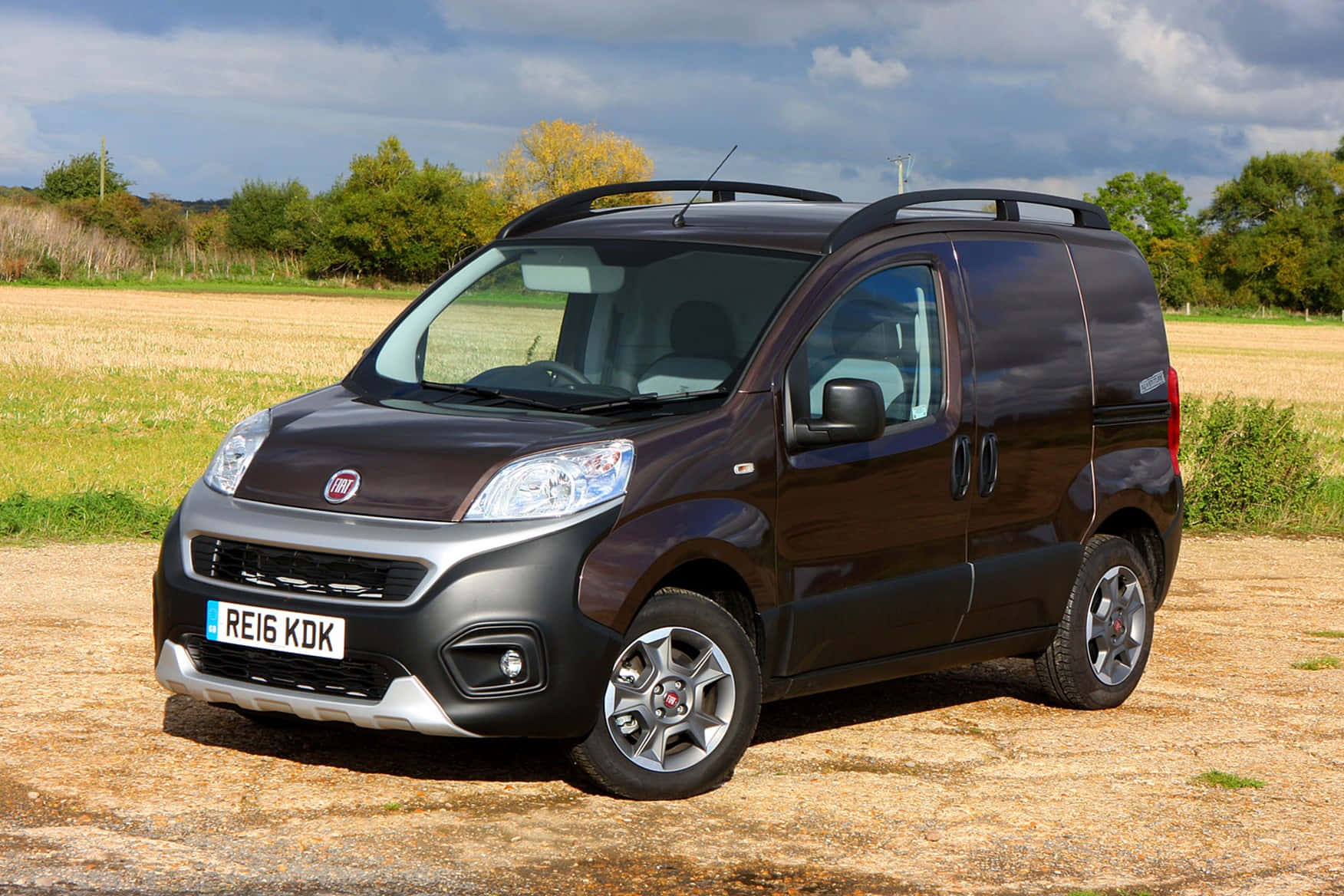 Fiat Fiorino 桌布 壁紙 — 一輛深棕色的Fiat廂型車停在碎石場上，天空多雲，背景有綠色和黃色的樹木。