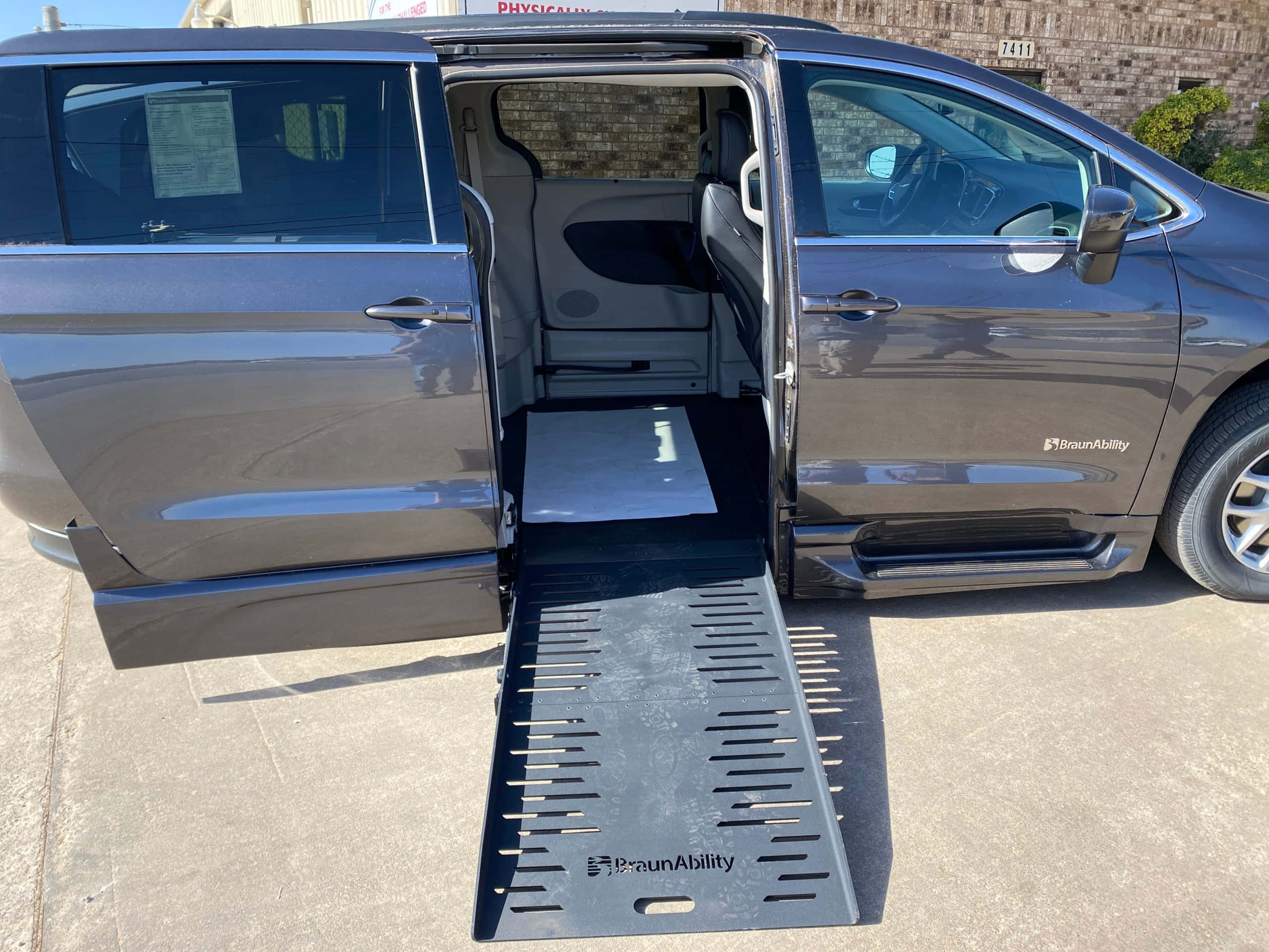 Chrysler Voyager 桌布 壁紙 — 一輛深灰色的BraunAbility廂型車，側門打開，金屬斜坡延伸到混凝土車道上，在明亮的陽光下。