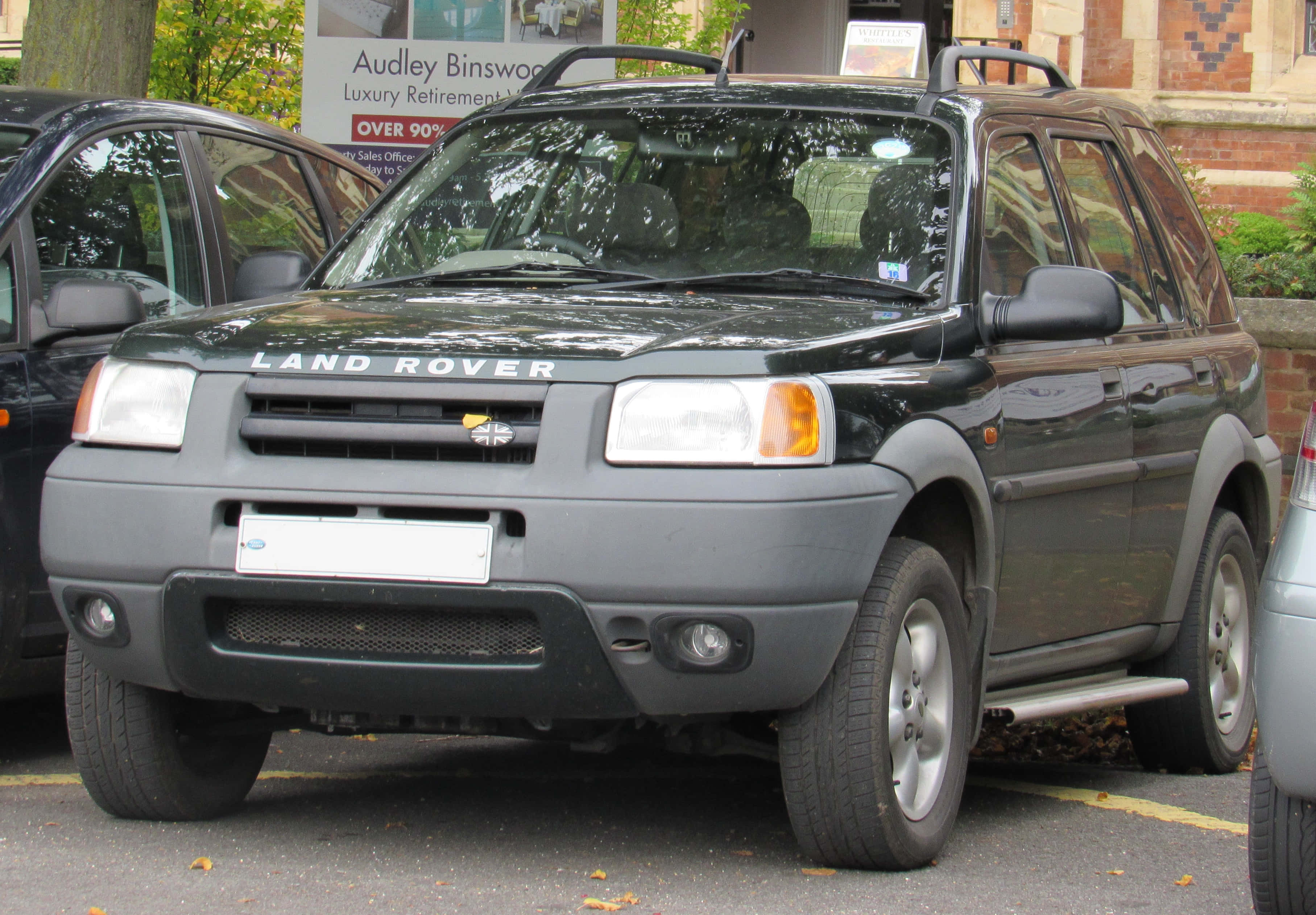 Land Rover Freelander 桌布 壁紙 — 一輛深灰色的 Land Rover Freelander 停在落葉遍地的城市空地，旁邊有磚造建築和其他汽車。