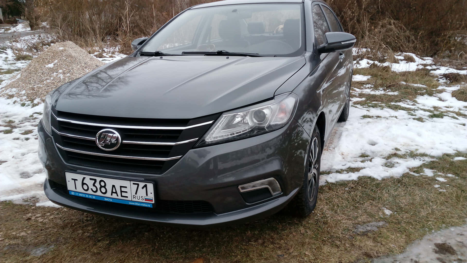  壁紙 — 一輛深灰色轎車停在積雪草地上，背景有光禿禿的灌木叢。
