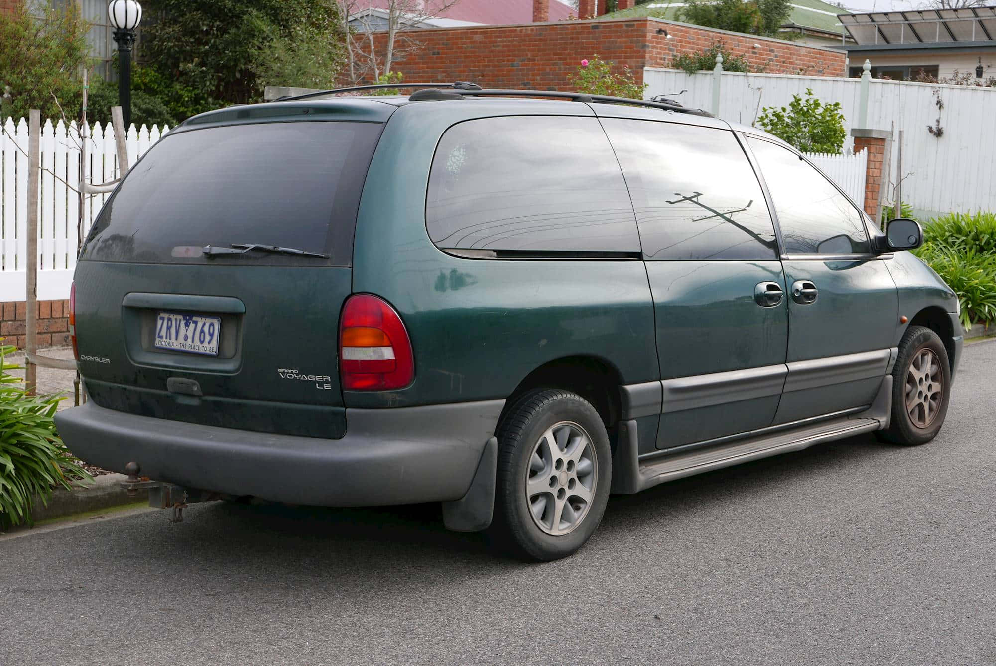 Chrysler Voyager 桌布 壁紙 — 一輛深綠色的克萊斯勒Voyager廂型車停在住宅區街道旁，旁邊有白色柵欄和磚牆。