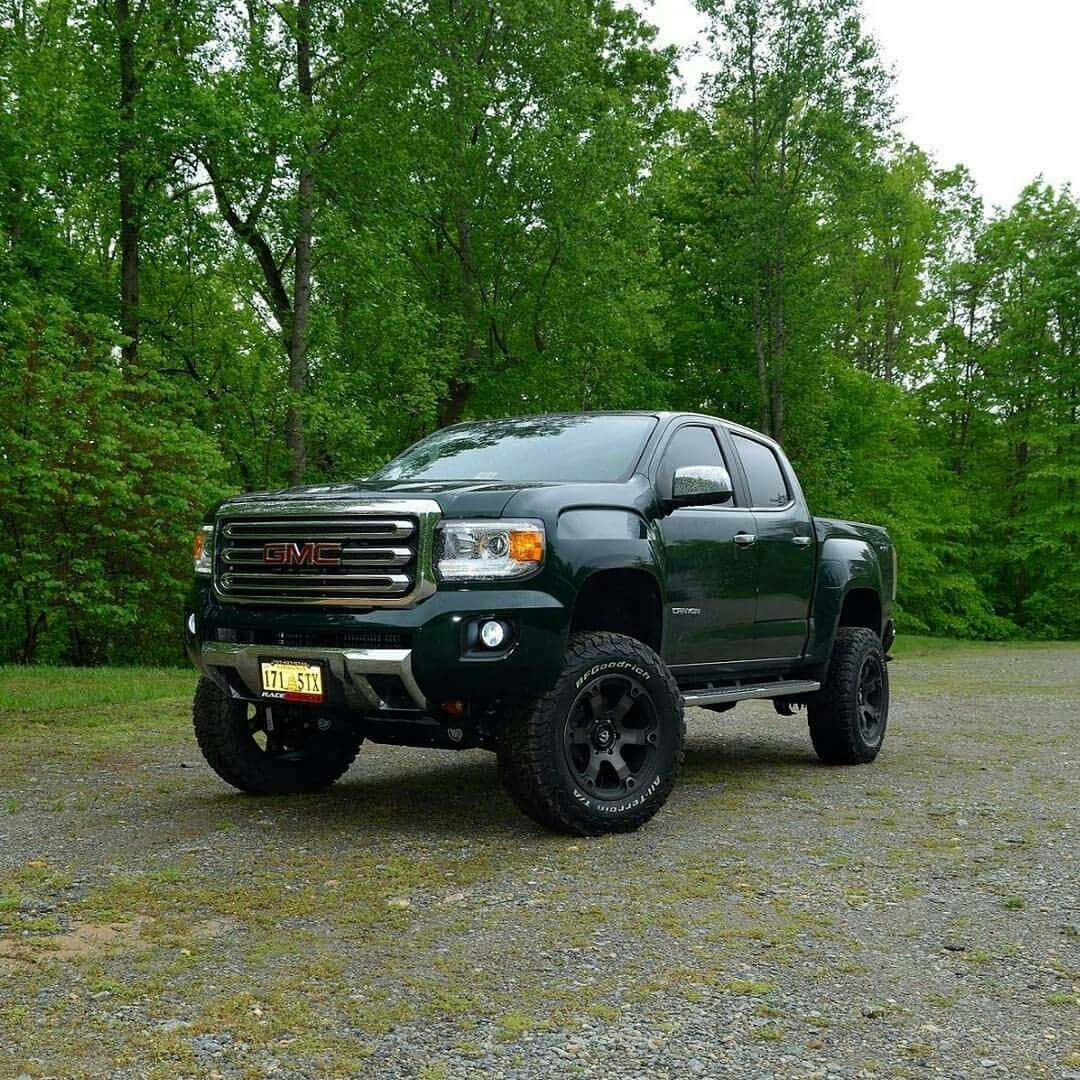 Gmc Canyon 桌布 壁紙 — 一輛深綠色GMC Sierra皮卡車，配備升高懸吊和黑色輪圈，停在碎石地上，周圍是茂密的綠樹。