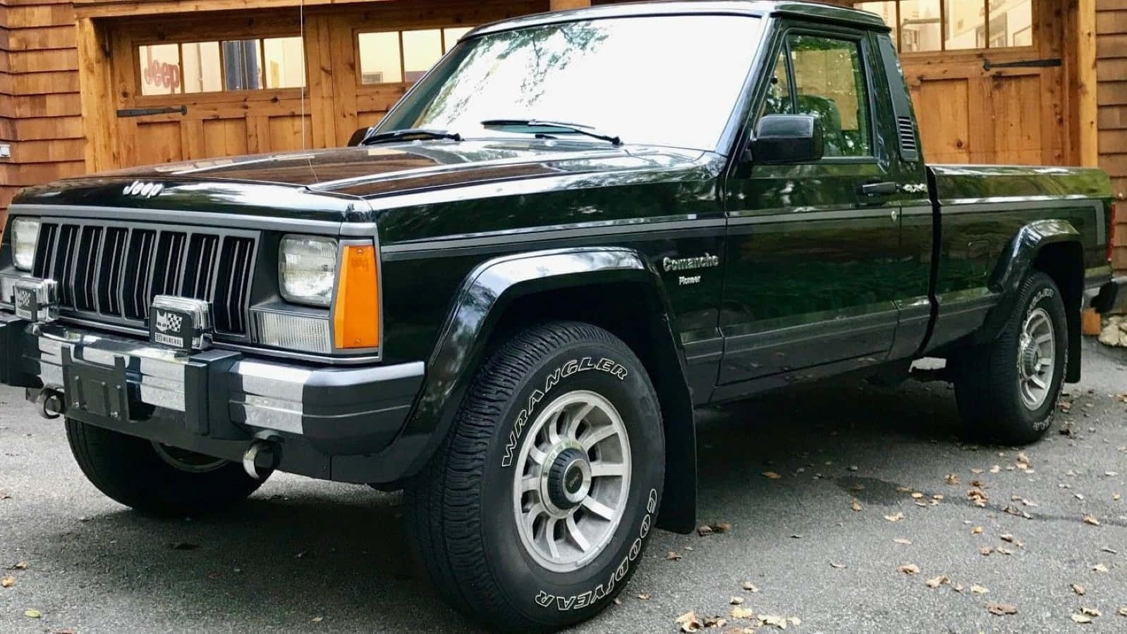 Jeep Comanche 桌布 壁紙 — 一輛深綠色Jeep Comanche皮卡車停在碎石路上，前面是木製車庫，周圍散落著落葉。