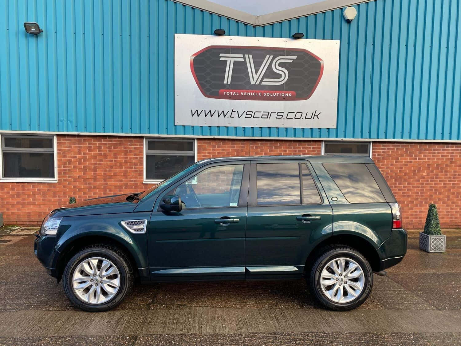 Land Rover Freelander 桌布 壁紙 — 一輛深綠色Land Rover Freelander SUV停在一棟藍色磚砌建築前，帶有TVS標誌。