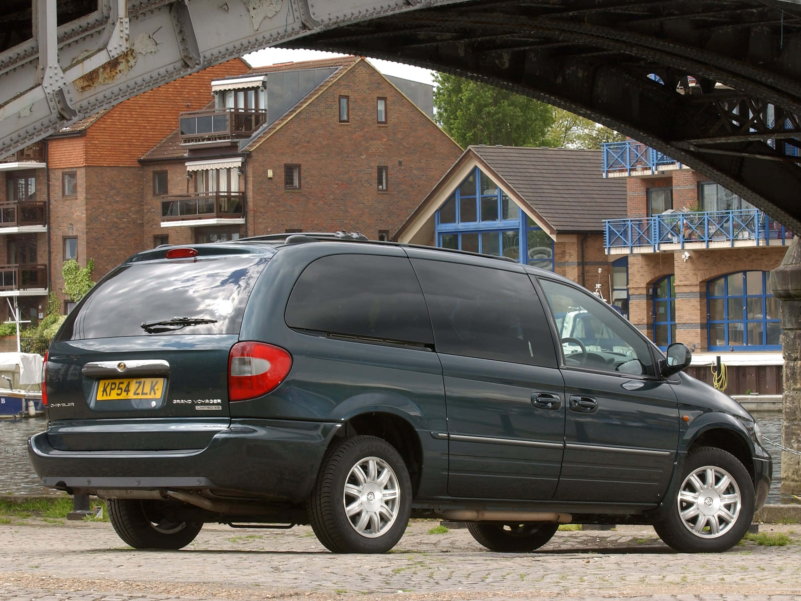 Chrysler Voyager 桌布 壁紙 — 一輛深綠色小型貨車停在鋼橋下的鵝卵石路上，背景有磚造建築和藍色陽台。