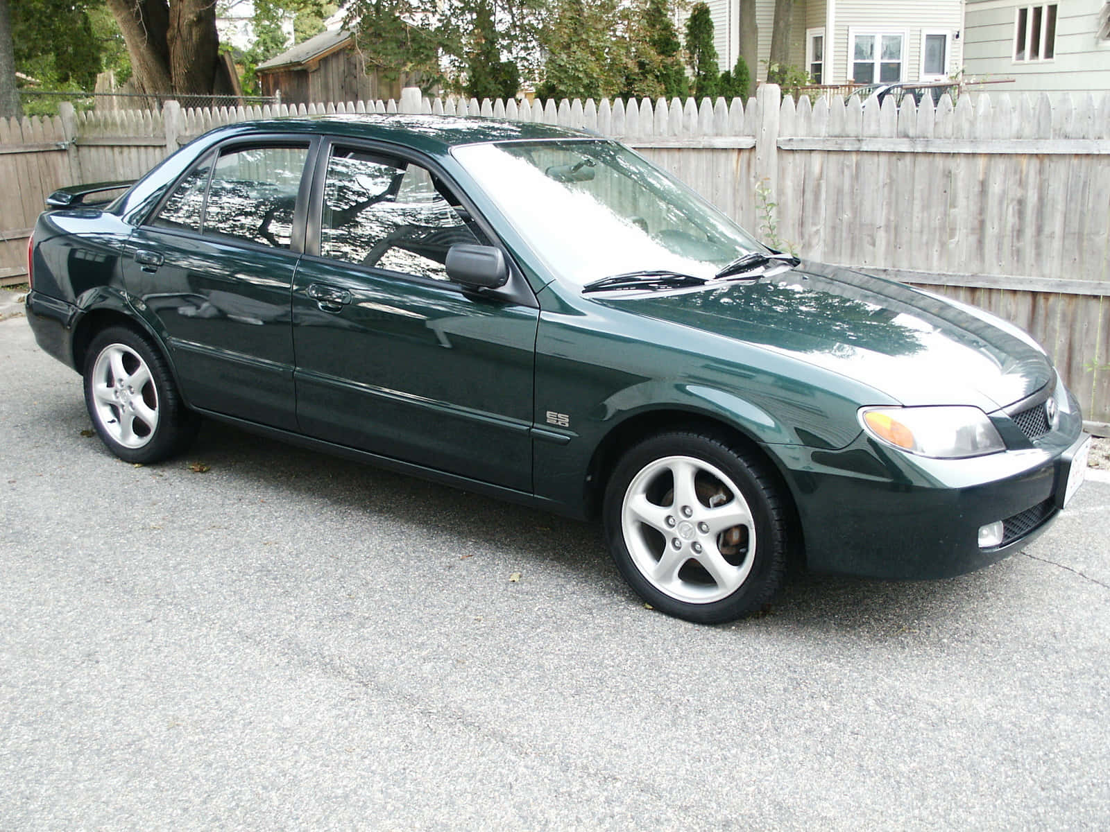 Mazda Protege 桌布 壁紙 — 一輛深綠色轎車配銀色輪圈，停在風化木柵欄前的碎石地上。