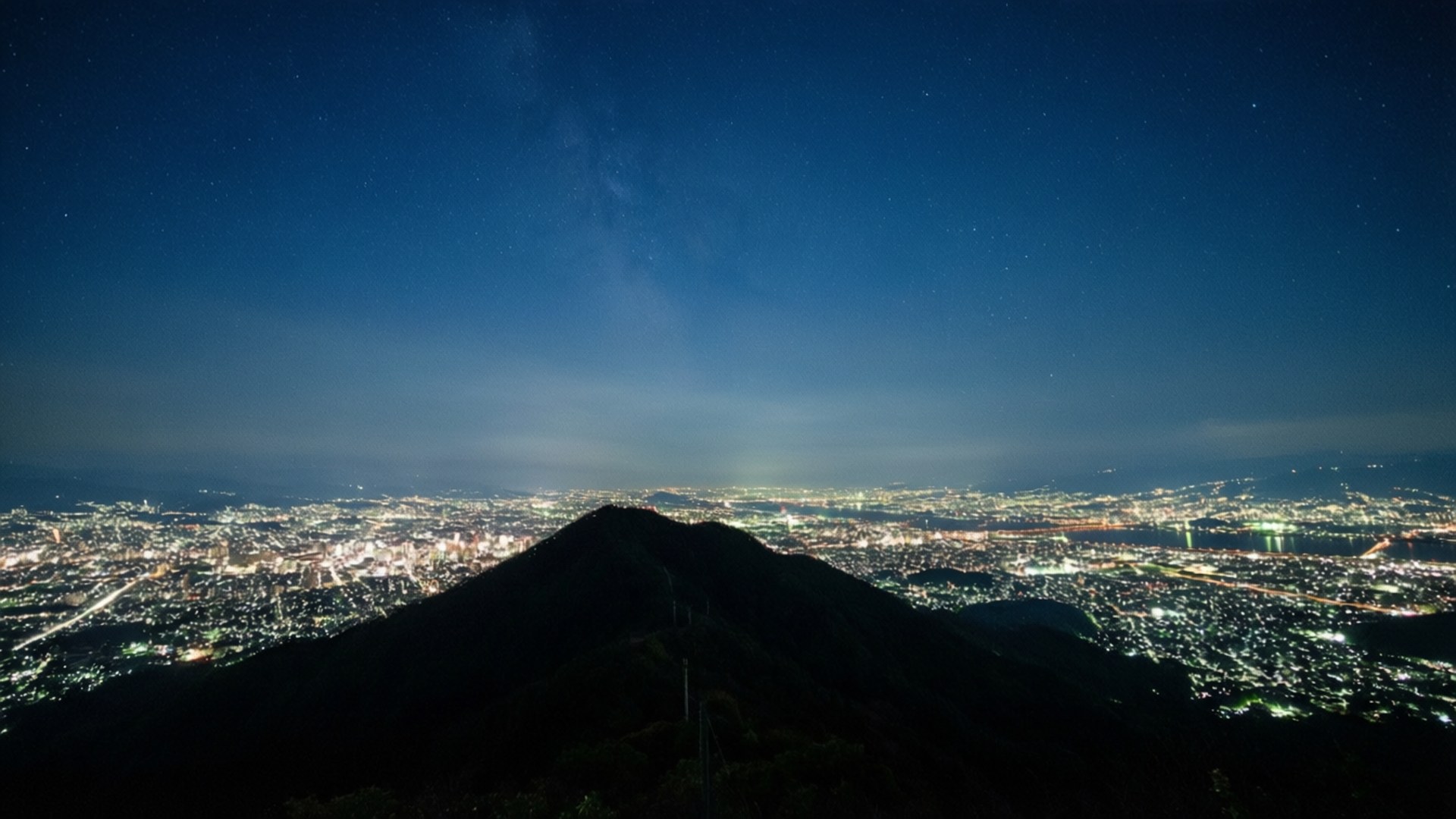 Mountain City Lights 壁紙 — 黑暗的山峰俯瞰著廣闊的城市景觀，在繁星點點的夜空下，銀河清晰可見，城市燈火輝煌。