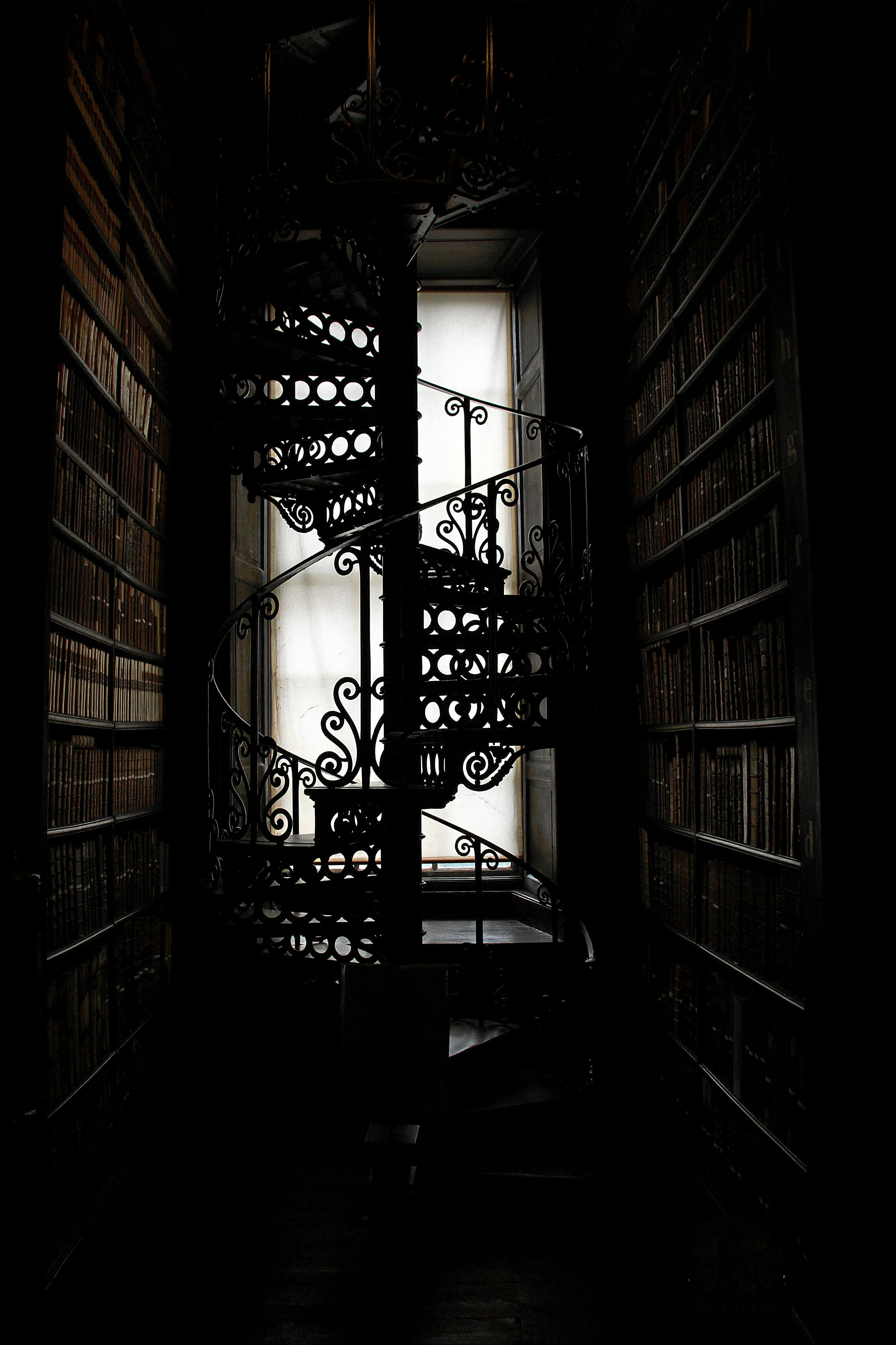 Dark Book Aesthetic 桌布 壁紙 — 一條深色華麗的螺旋樓梯穿過兩旁高聳書架的圖書館，通往明亮的窗戶。