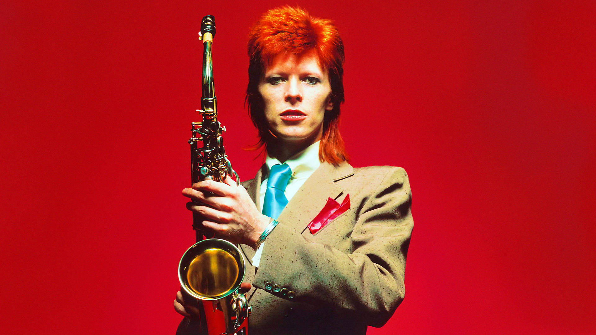 大衛·鮑伊 桌布 壁紙 — David Bowie 一頭火紅頭髮，身穿藍色領帶的俐落西裝，在鮮紅色背景下拿著薩克斯風。