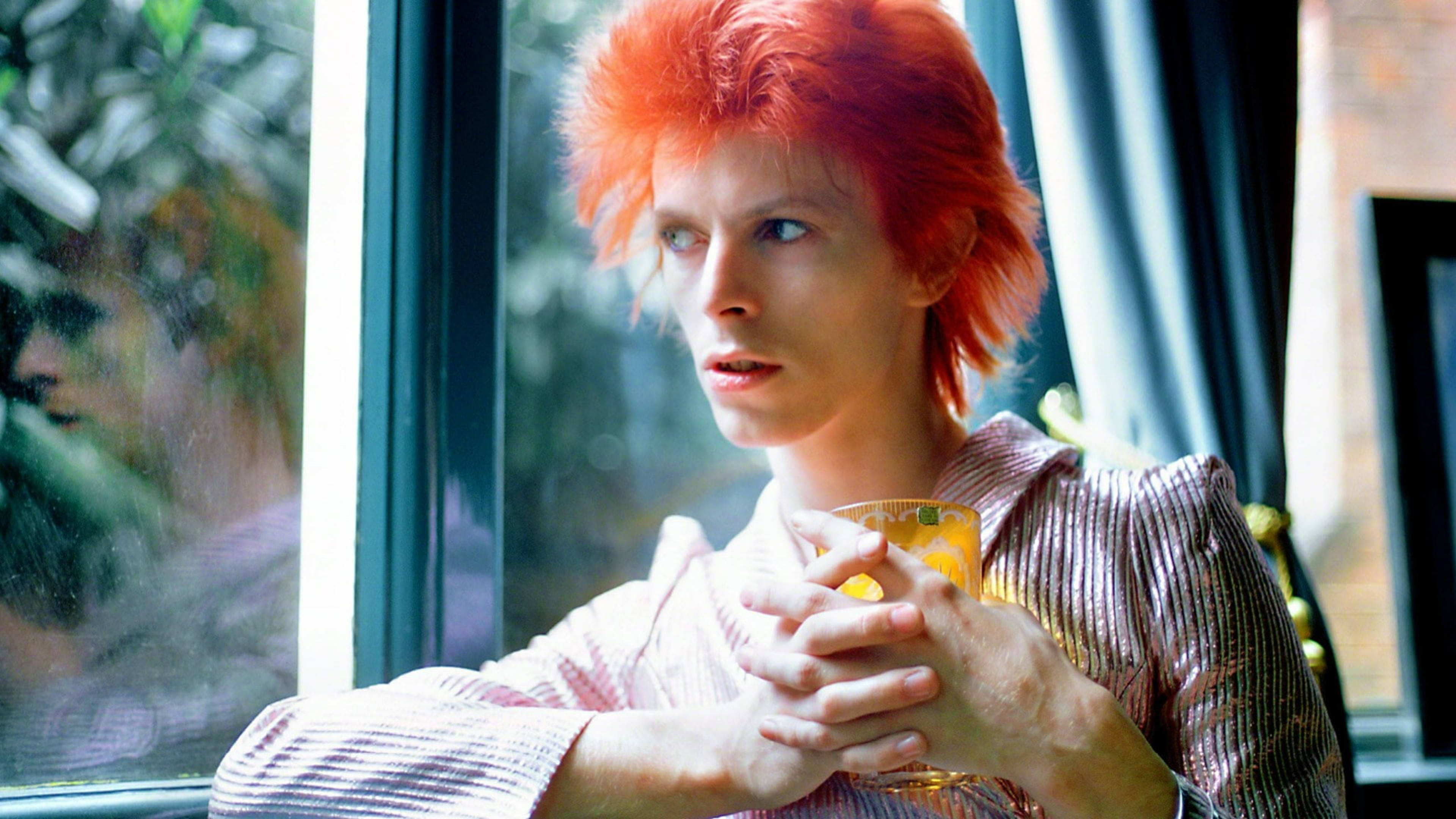 大衛·鮑伊 桌布 壁紙 — David Bowie 一頭火紅頭髮，身穿條紋襯衫，在陽光照射的窗邊拿著飲品。