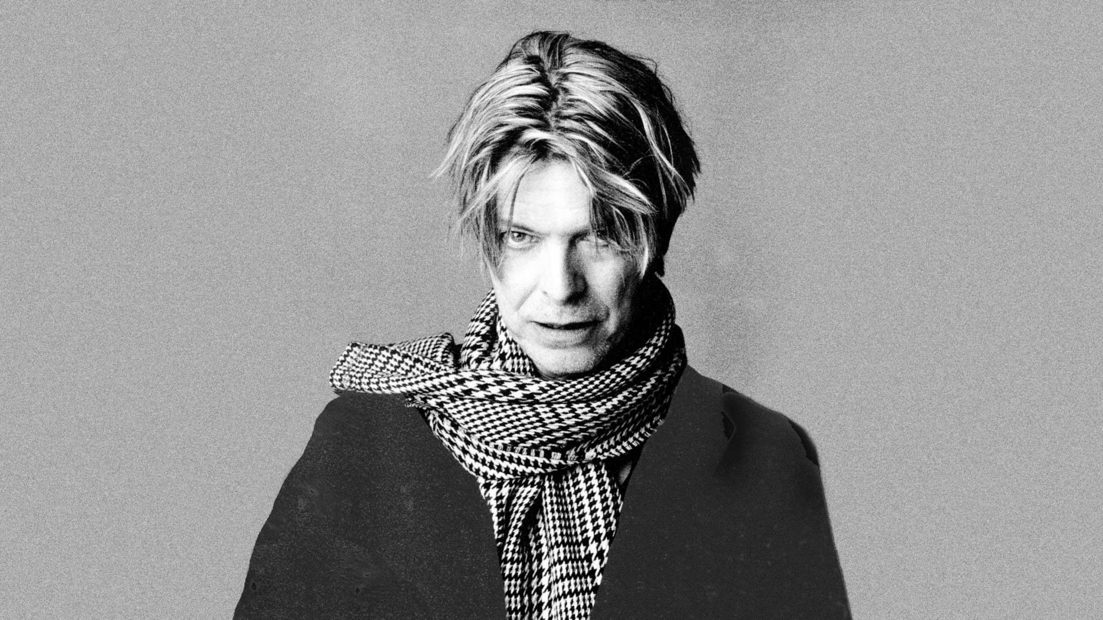 大衛·鮑伊 桌布 壁紙 — David Bowie 在黑白肖像中強烈凝視，穿著千鳥格圍巾，背景為純灰色。