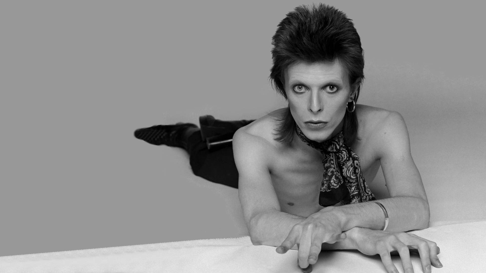 大衛·鮑伊 桌布 壁紙 — David Bowie 赤裸上身躺在白色表面上，帶著戲劇性的化妝和圖案圍巾，強烈地凝視著相機。