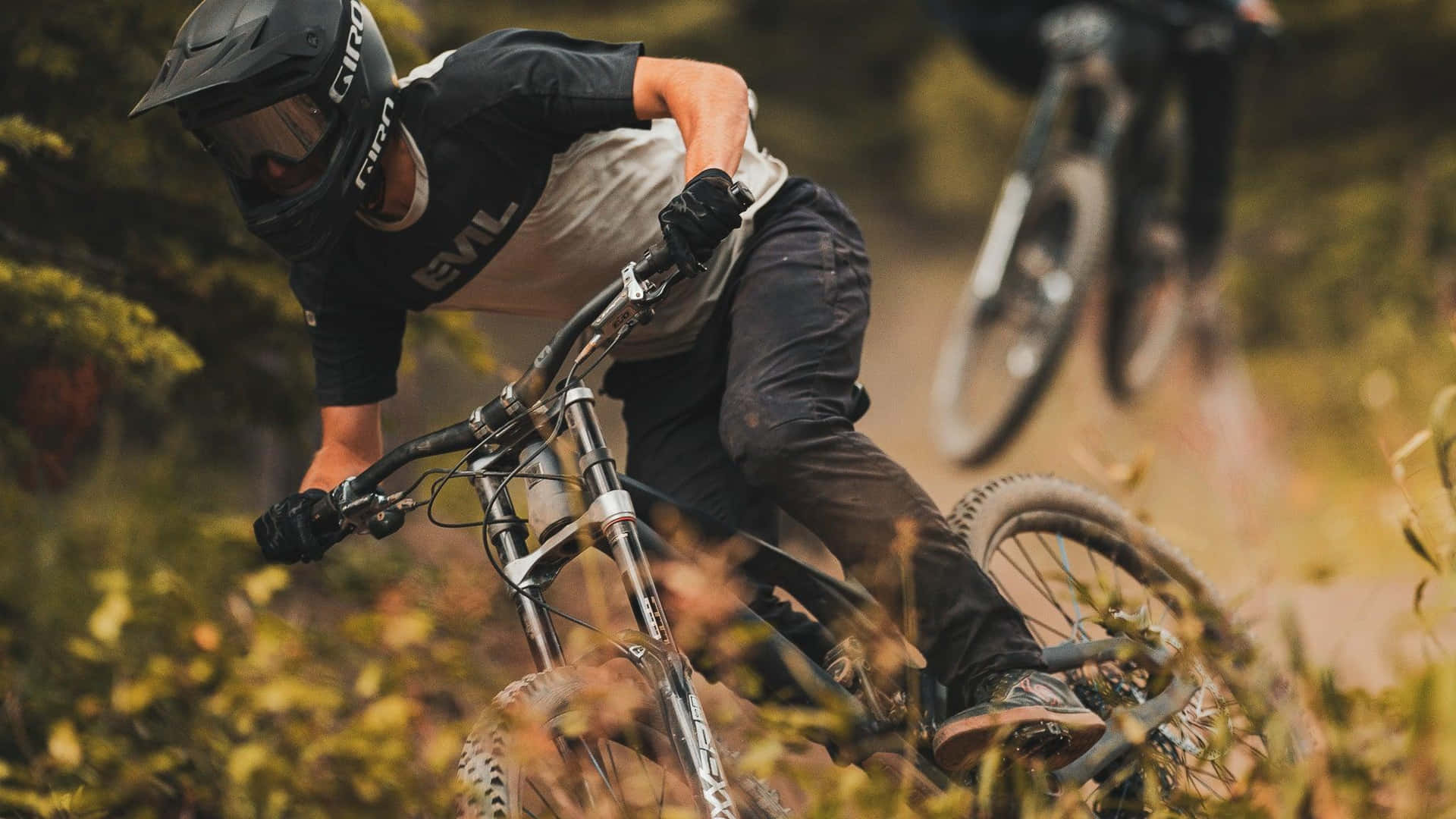  壁紙 — 一名登山車騎士在森林小徑上傾斜進入一個急彎，揚起灰塵和草，背景中另一名騎士模糊不清。