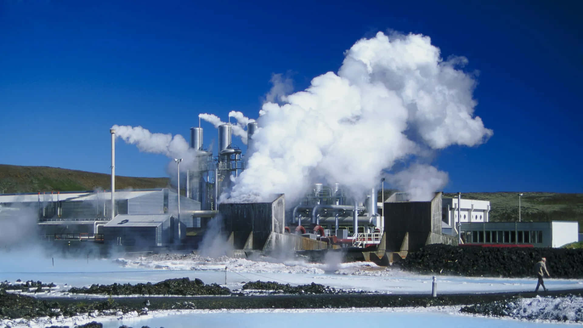  壁紙 — 一座地熱發電廠在晴朗的藍天下噴出濃厚的白色蒸氣，附近有一個孤獨的人走過。
