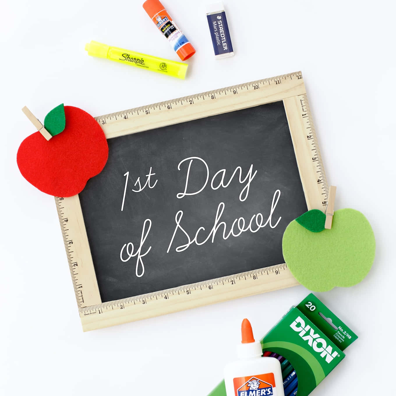  壁紙 — 一個木框黑板，上面用白色草書寫著「1st Day of School」，兩側有氈蘋果，周圍是白色背景上的學校用品。