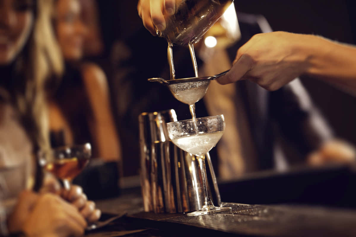  壁紙 — 調酒師將冰涼雞尾酒透過濾網倒入碟形杯，而顧客在昏暗的酒吧中手持飲料。