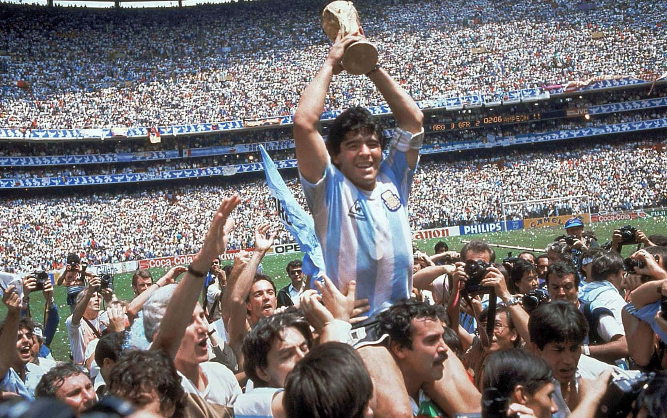 足球 桌布 壁紙 — Diego Maradona 高舉世界盃獎盃在頭頂，歡欣鼓舞的隊友和球迷在陽光普照的球場上慶祝。