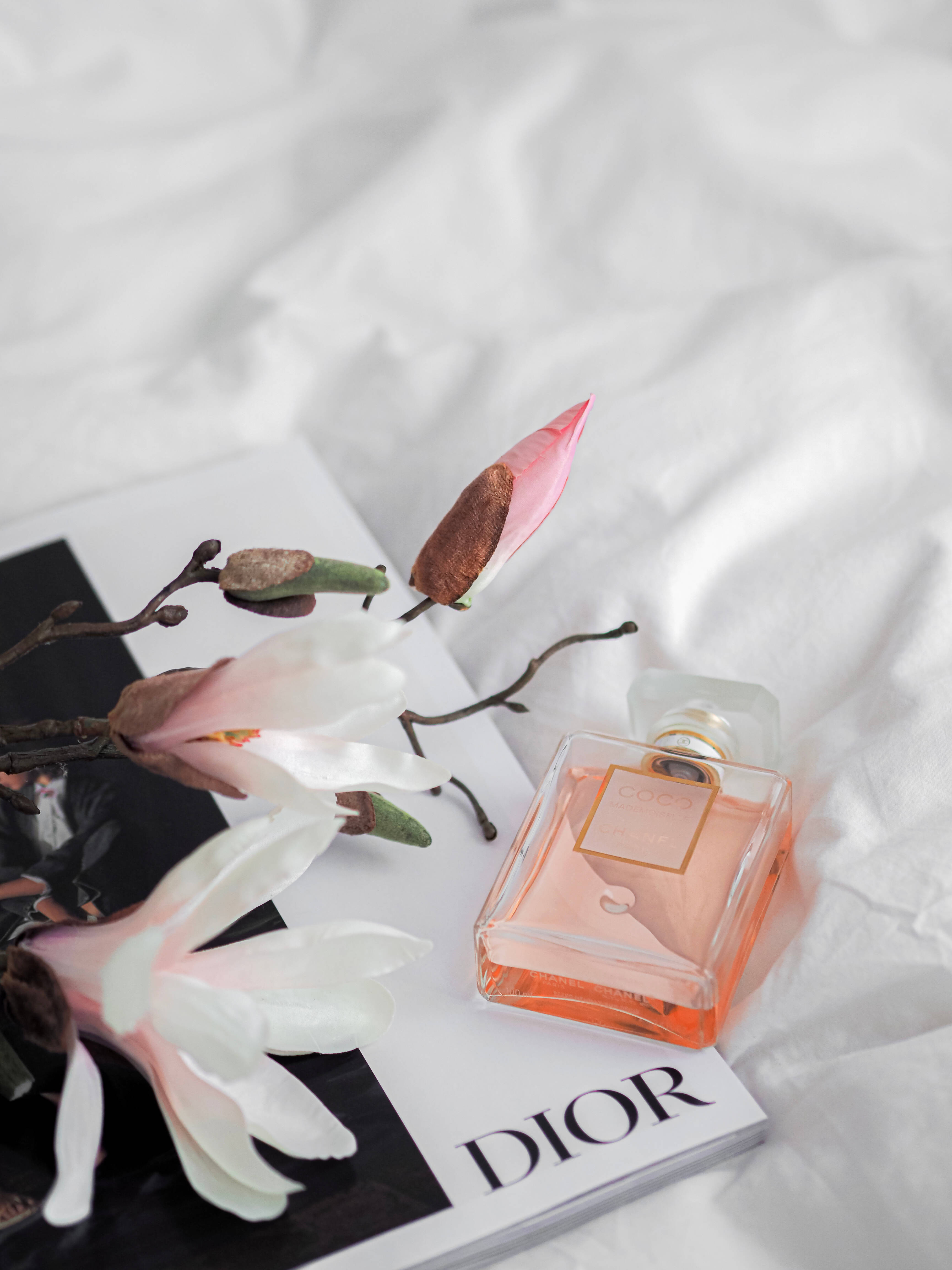 Dior 動態 桌布 壁紙 — Dior香水瓶放在白色雜誌上，旁邊有木蘭花，底下是皺巴巴的床單。