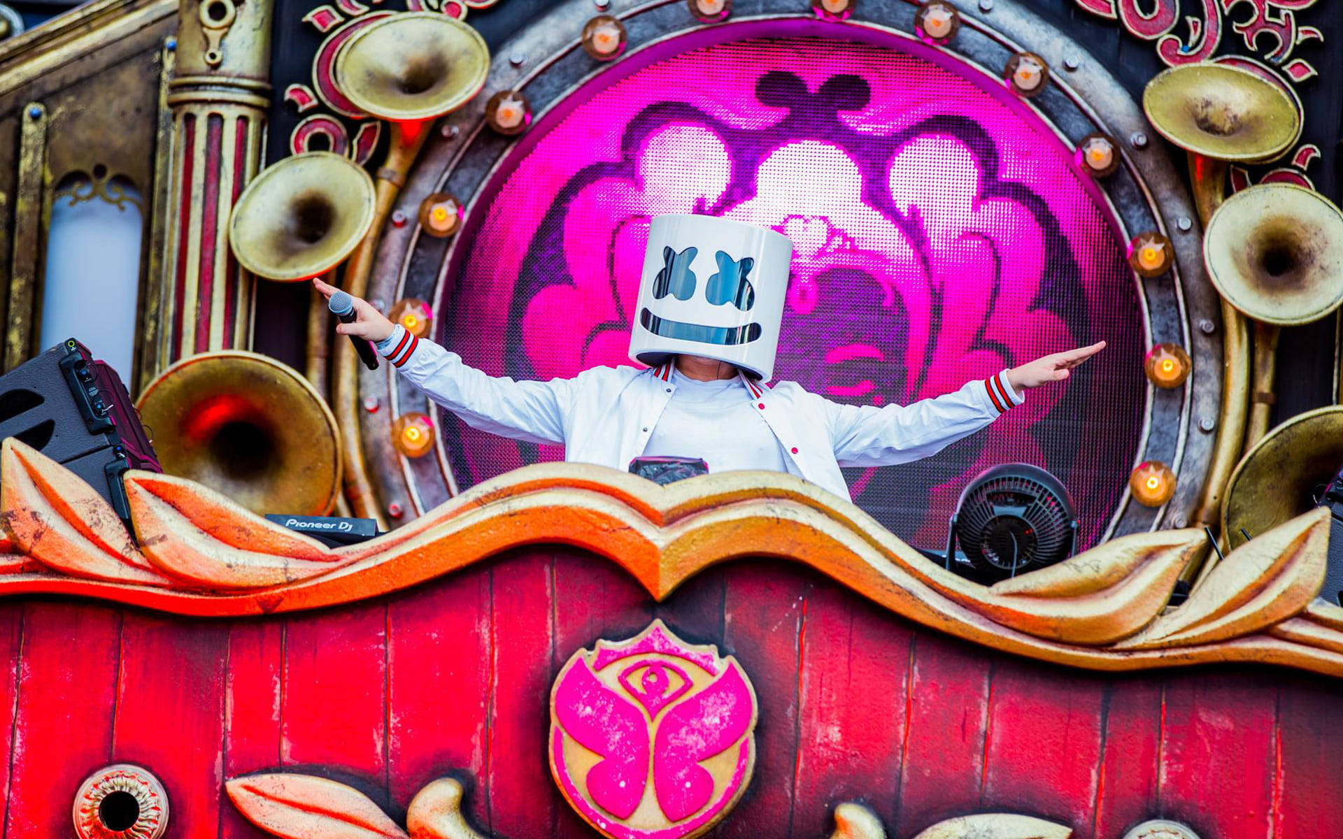 明日世界 桌布 壁紙 — DJ Marshmello 在一個充滿活力的紅色和金色舞台上表演，身後有華麗的黃銅喇叭和發光的粉紅色螢幕。