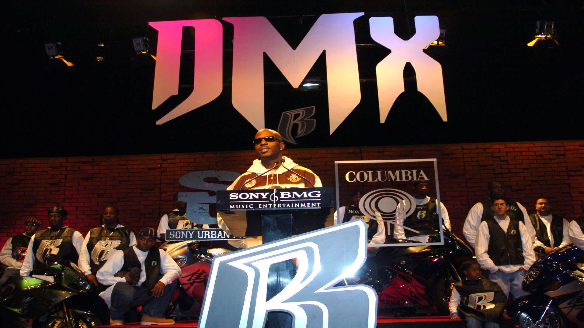 DMX 桌布 壁紙 — DMX 自信地站在講台上，身後是一個巨大的發光 'DMX' 標誌，他的團隊騎著摩托車在兩側護航，活動空間燈光昏暗。