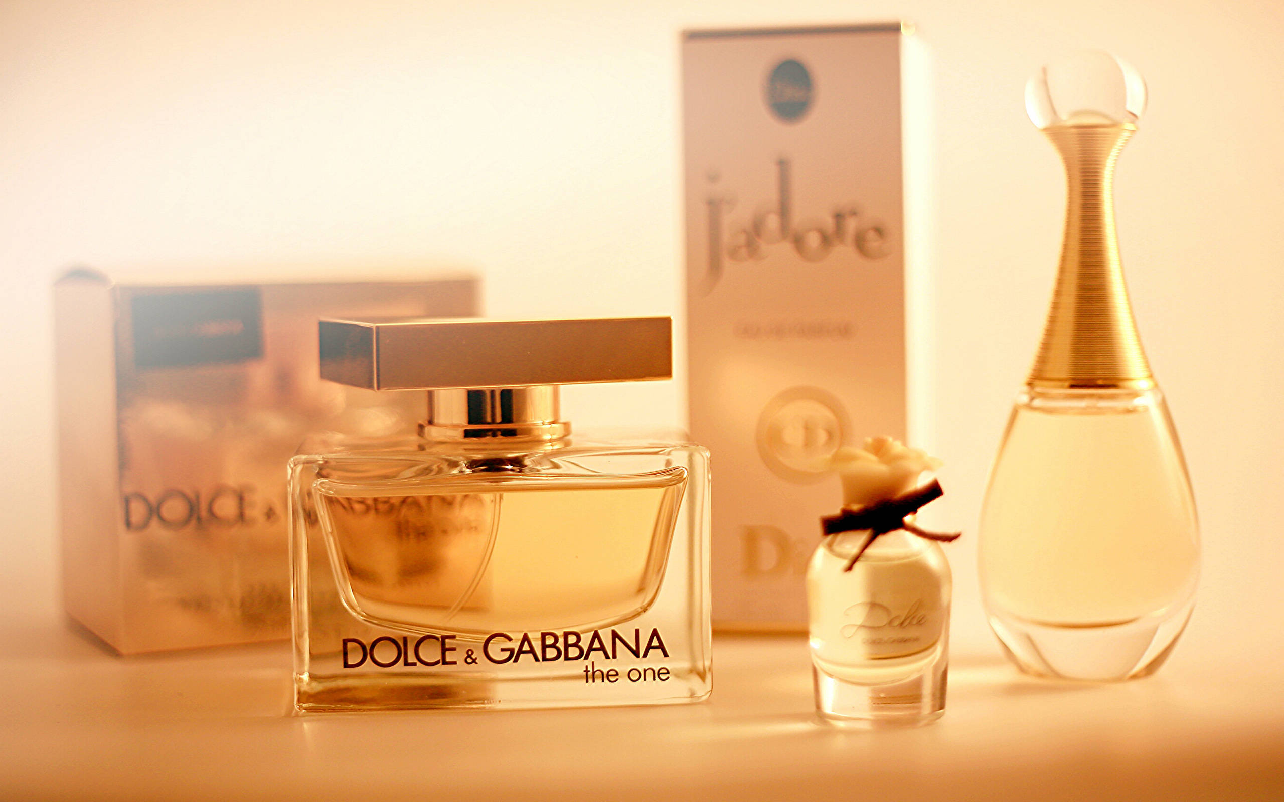 時尚 桌布 壁紙 — Dolce & Gabbana 和 Dior 的金色香水瓶和盒子排列在溫暖的照明表面上。