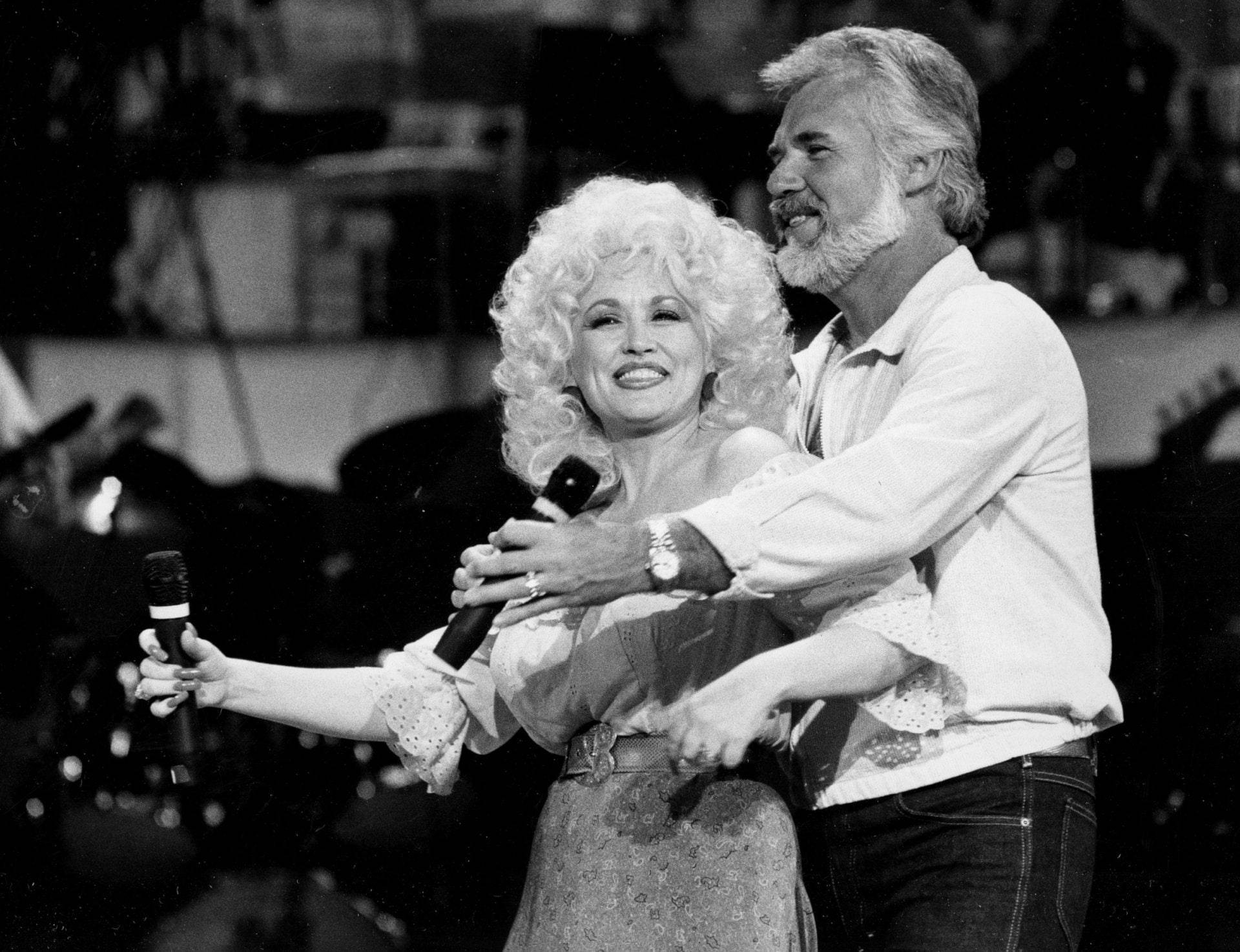 dolly 桌布 壁紙 — Dolly Parton 和 Kenny Rogers 在現場表演中手持麥克風在舞台上歡樂擁抱。