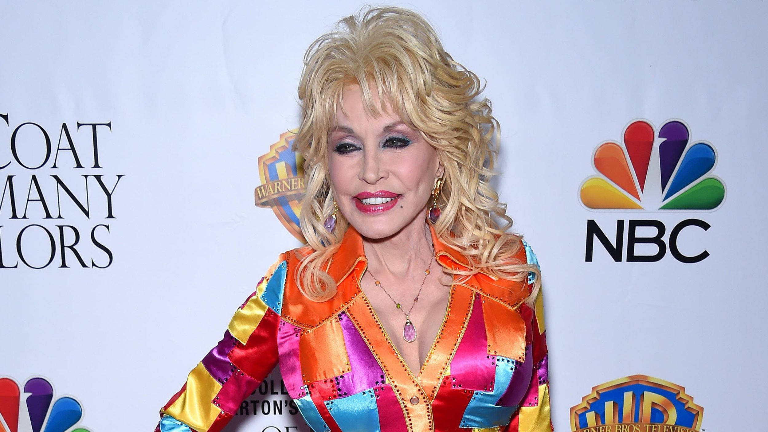 dolly 桌布 壁紙 — Dolly Parton 在一個媒體活動中，身後有NBC和Warner Bros標誌，穿著鮮豔的拼布緞面外套，燦爛地微笑。