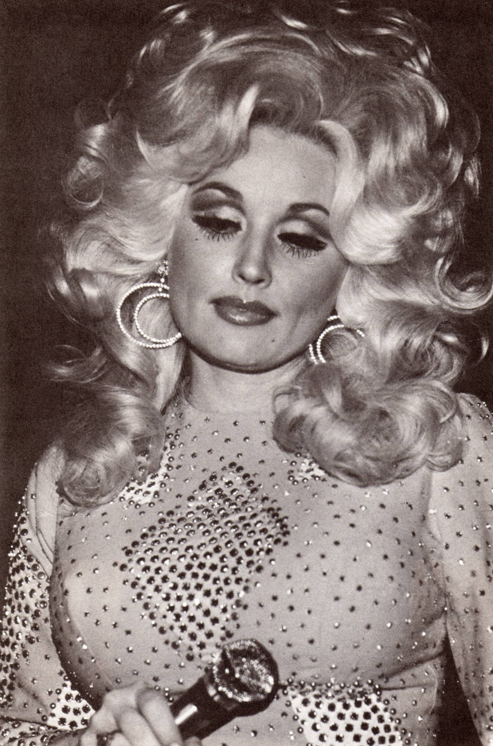 dolly 桌布 壁紙 — Dolly Parton 身穿閃亮水鑽服裝，手持麥克風，擁有豐盈的金色捲髮和大圈耳環。