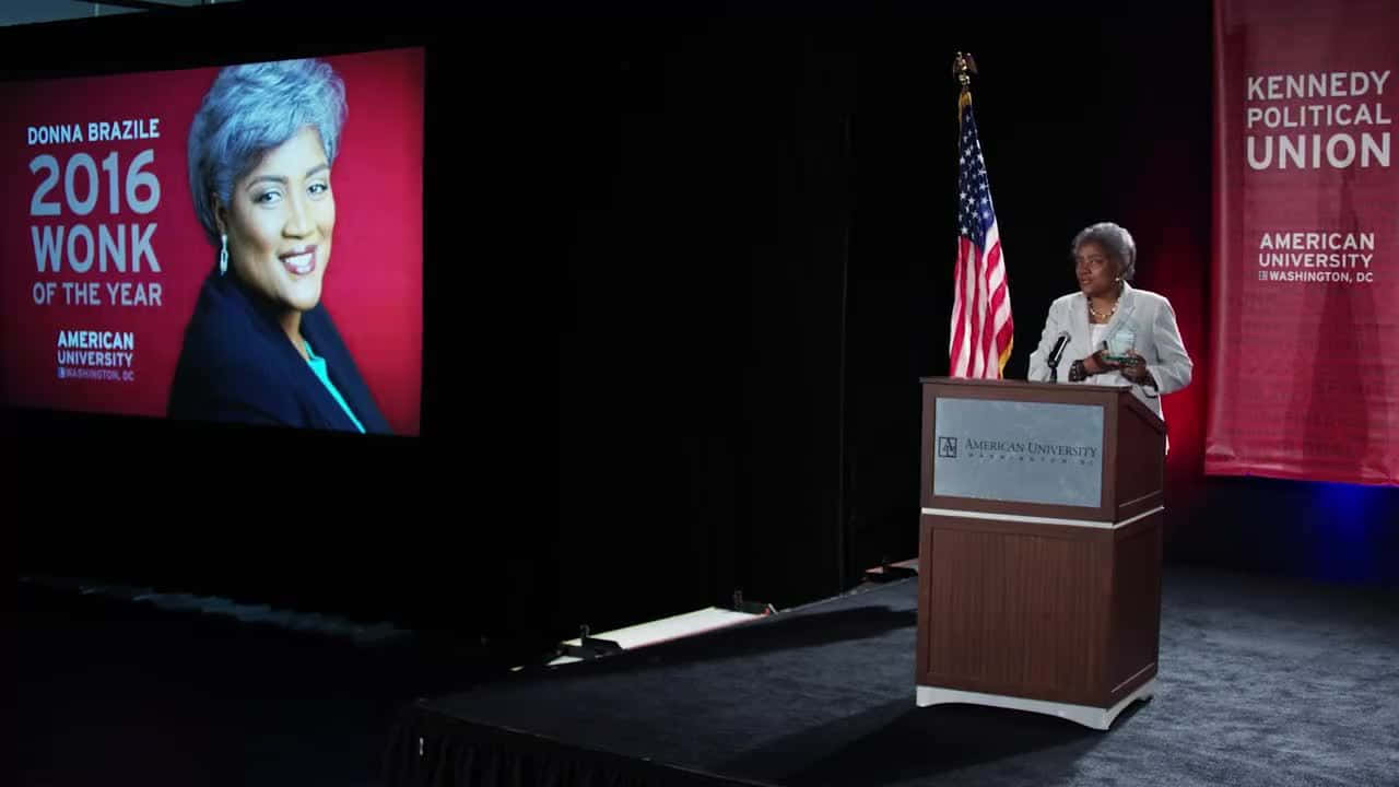 政治 桌布 壁紙 — Donna Brazile 站在燈光昏暗的舞台講台上，旁邊有美國國旗，身後的大螢幕顯示她的肖像以及美國大學頒發的「2016 Wonk of the Year」獎項。