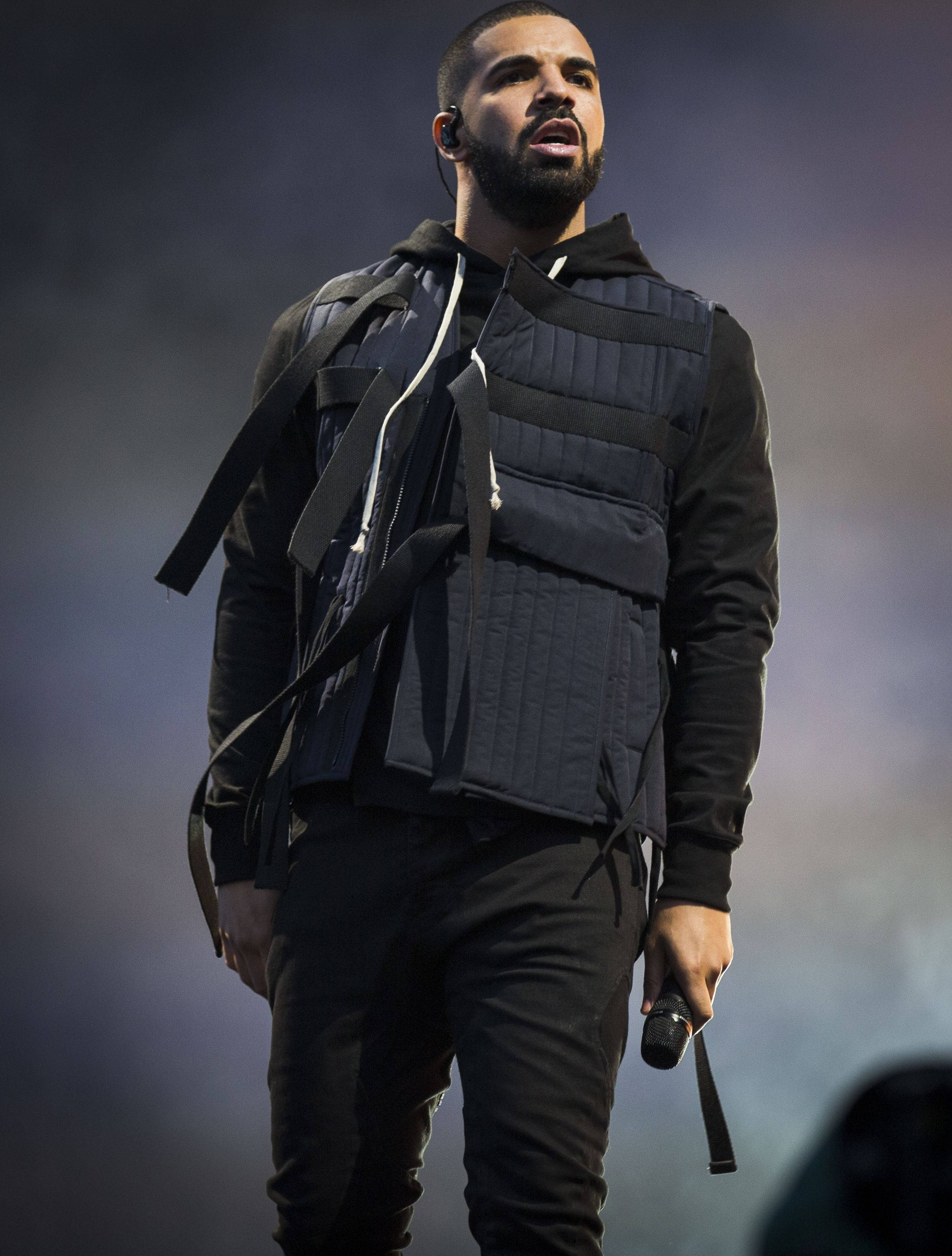 Drake 桌布 壁紙 — Drake 在戲劇性的舞台燈光下，穿著黑色絎縫背心、帶子和連帽衫在舞台上表演。