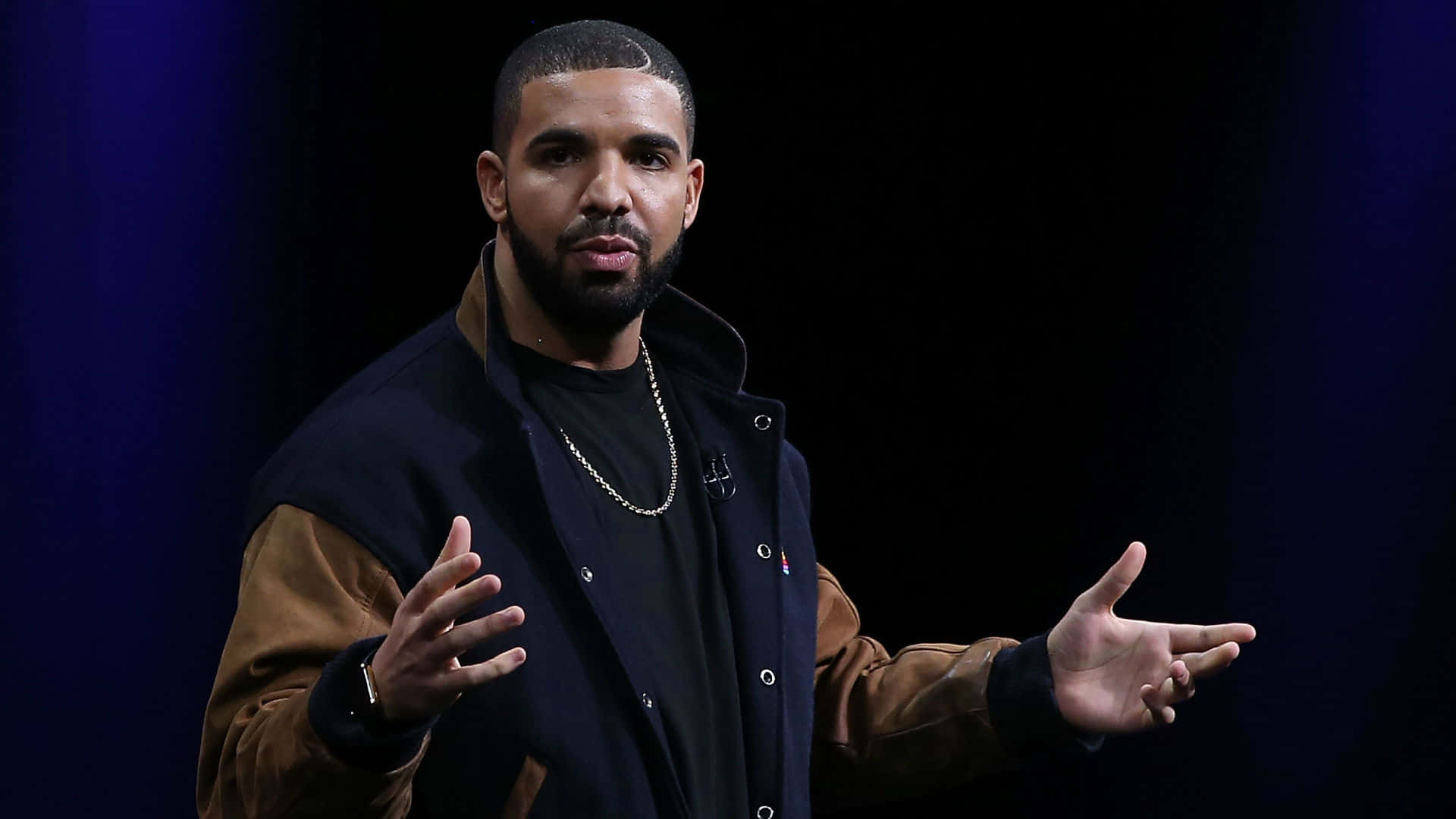 桌布 壁紙 — Drake 在深藍色背景前，自信地做手勢，穿著深藍色和棕色的外套。