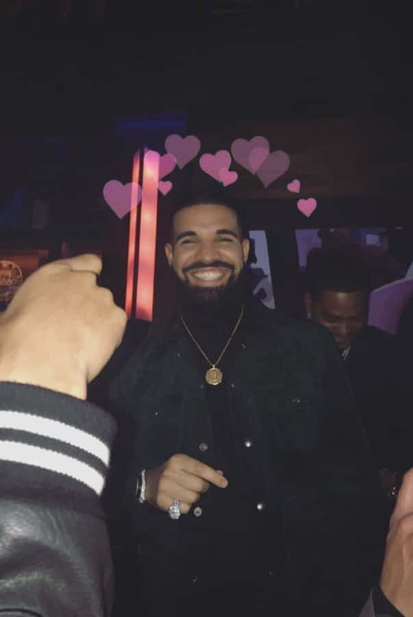 Aesthetic Drake 桌布 壁紙 — Drake 在燈光昏暗的派對上燦爛地微笑，粉紅色心形圖案漂浮在他的頭頂上方。