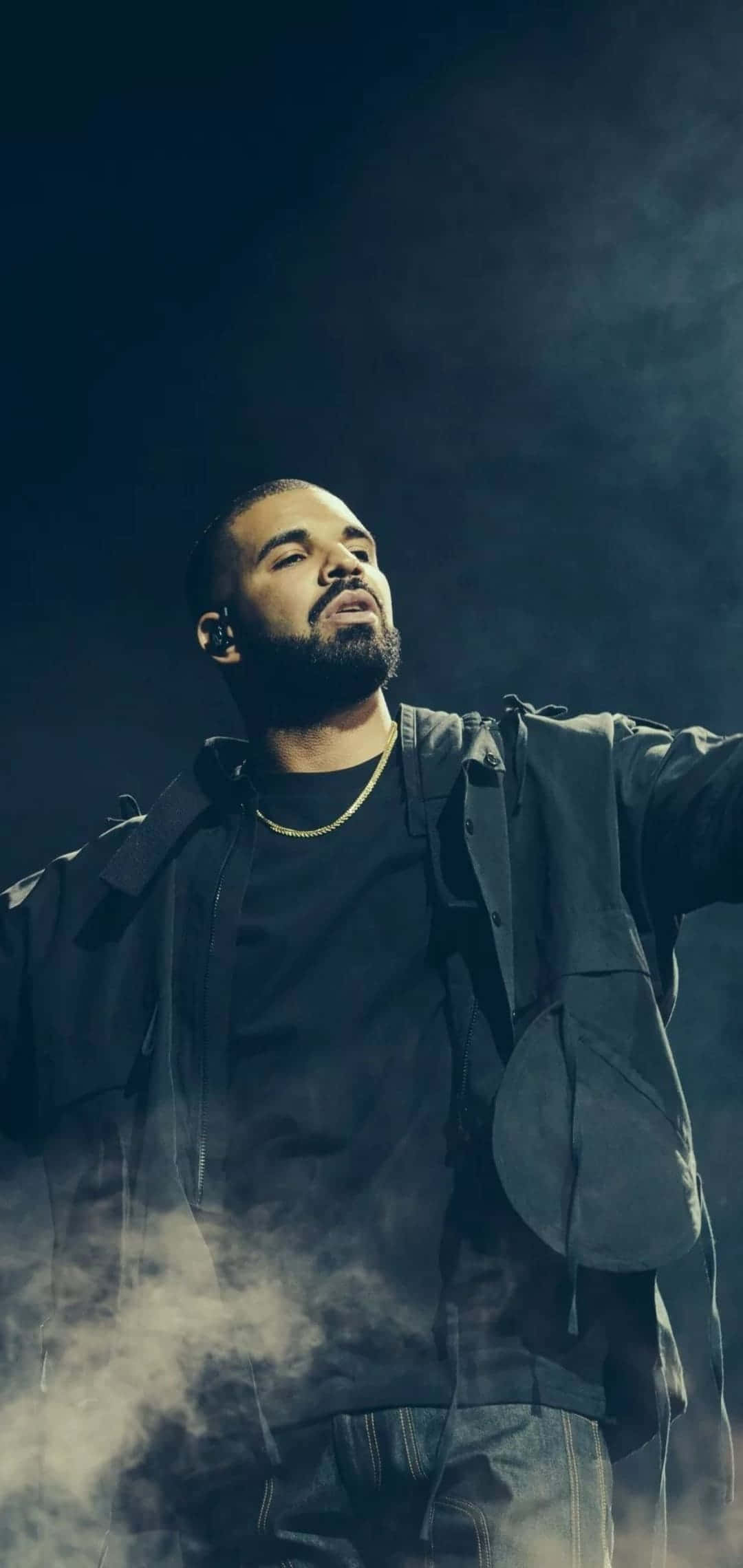 Aesthetic Drake 桌布 壁紙 — Drake 在舞台上表演，身穿深色外套和牛仔褲，煙霧在他周圍旋轉，燈光效果戲劇化。