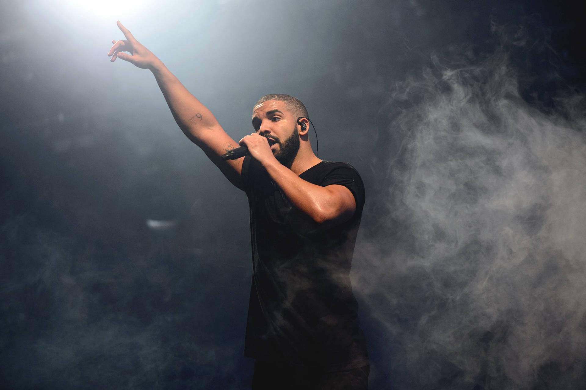 Drake 桌布 壁紙 — Drake 在黑暗煙霧中於明亮聚光燈下在舞台上表演，手持麥克風並向上指。