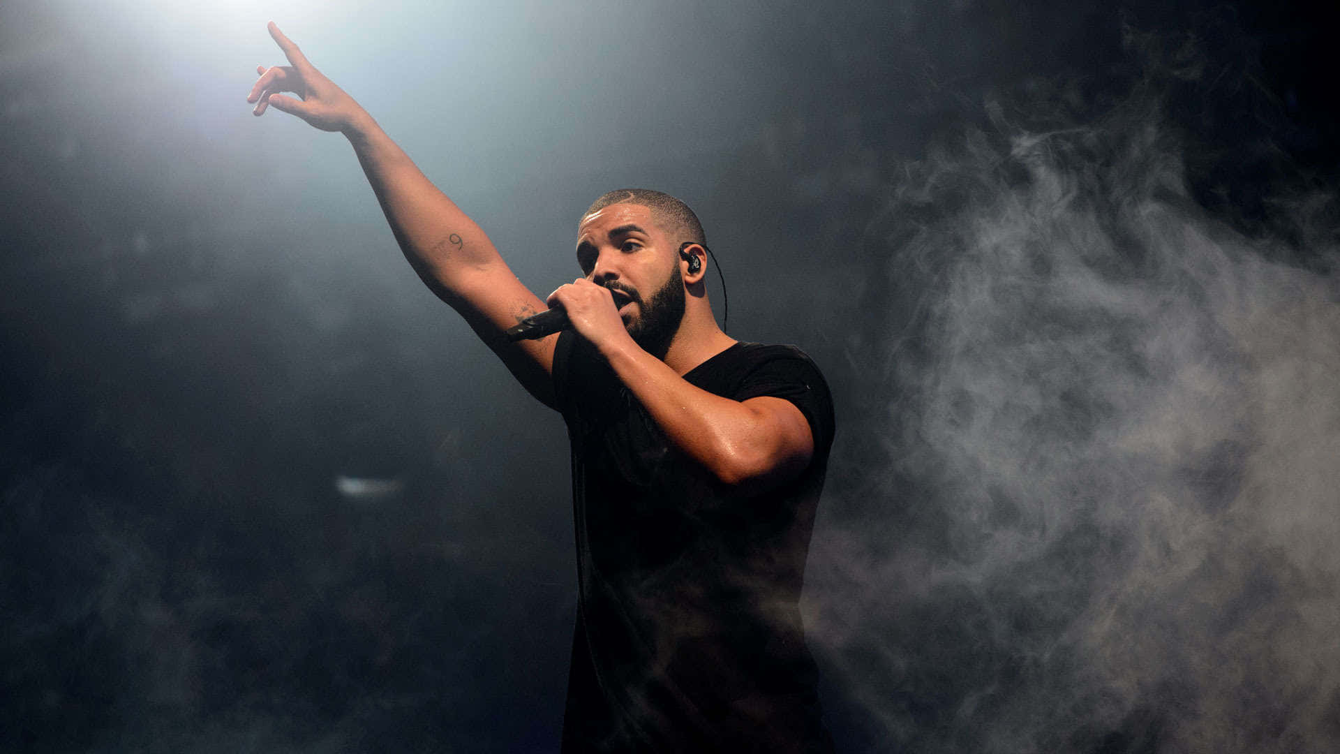 桌布 壁紙 — Drake 在黑暗煙霧中於明亮聚光燈下在舞台上表演，手持麥克風並向上指。
