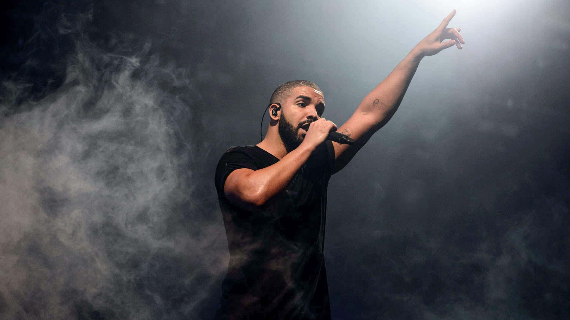 Drake 桌布 壁紙 — Drake 在黑暗煙霧中於舞台上表演，單手舉起，手持麥克風，在明亮的聚光燈下。