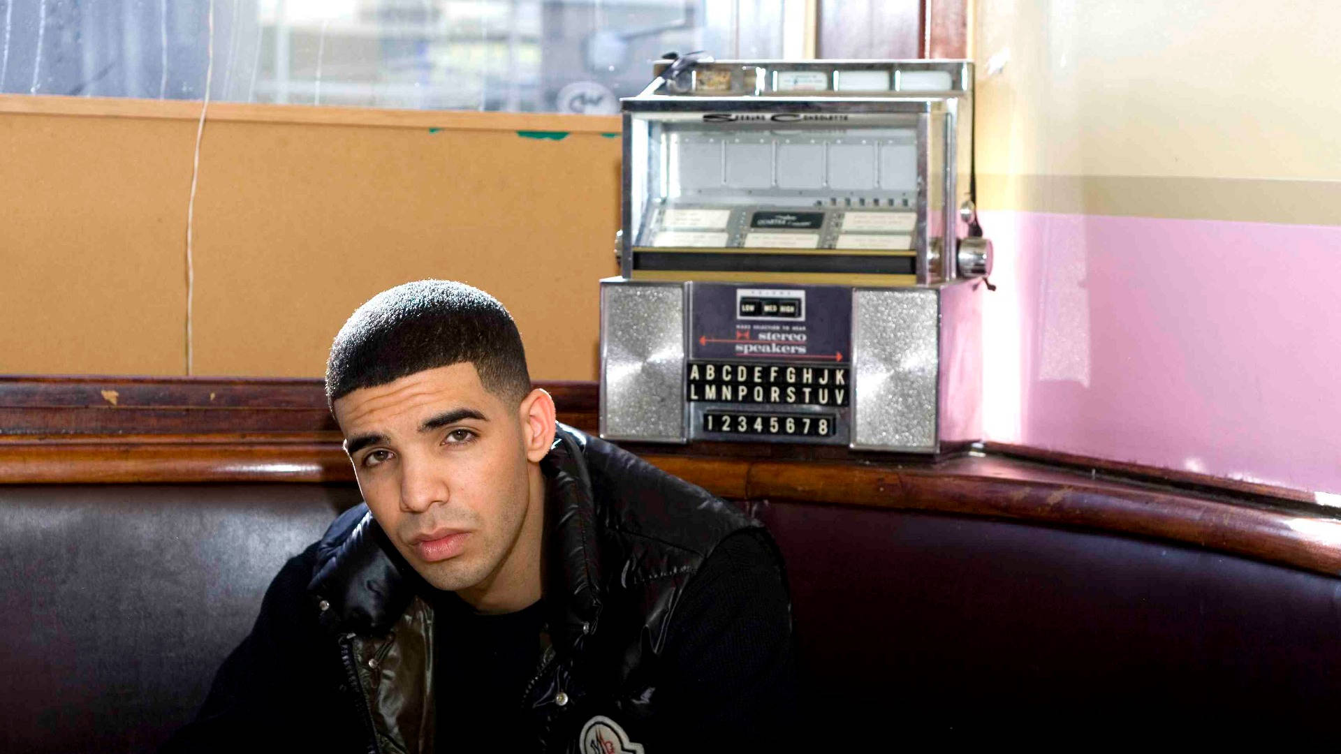 Drake 桌布 壁紙 — Drake 坐在復古餐廳卡座旁，旁邊有一台懷舊點唱機，身後是粉紅色和米色的牆壁。