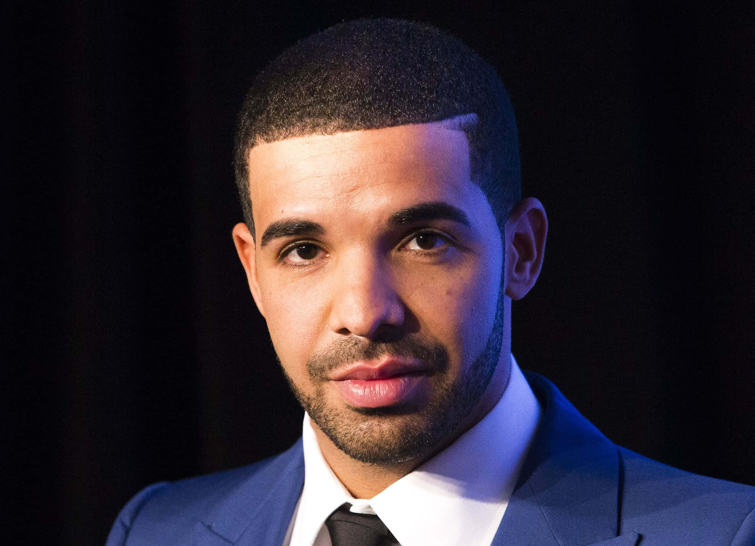 Drake 桌布 壁紙 — Drake 的特寫肖像，穿著一套俐落的藍色西裝，在黑暗背景前搭配戲劇性燈光。
