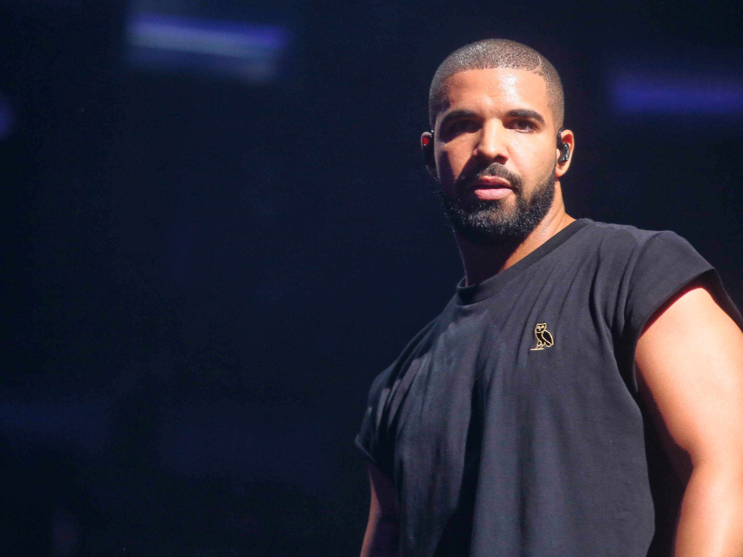 Drake 桌布 壁紙 — Drake 自信地站在舞台上，身穿黑色無袖襯衫，在戲劇性的藍色舞台燈光下。