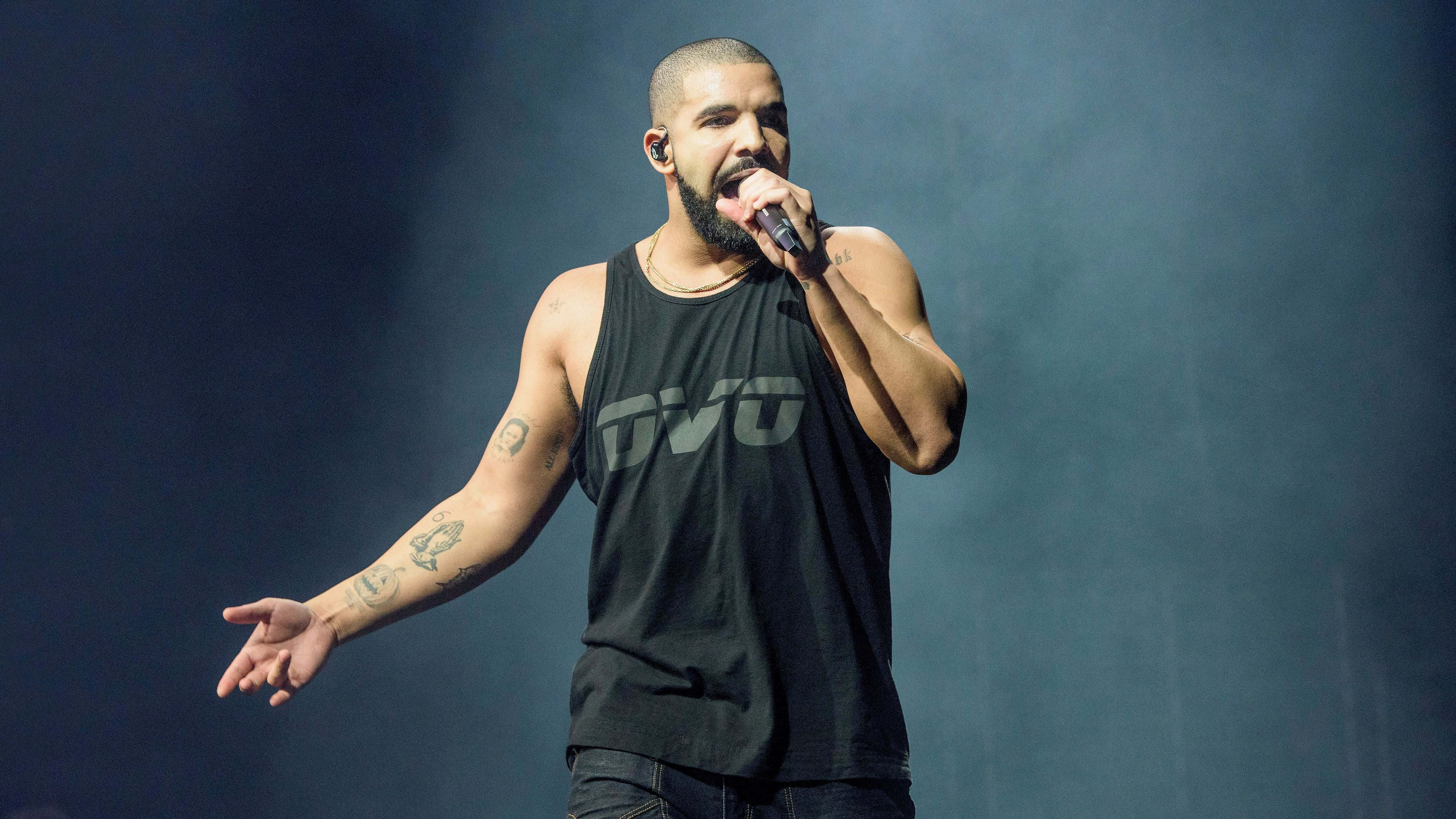 Drake 桌布 壁紙 — Drake 身穿黑色背心在舞台上，在戲劇性的藍色舞台燈光下手持麥克風表演。