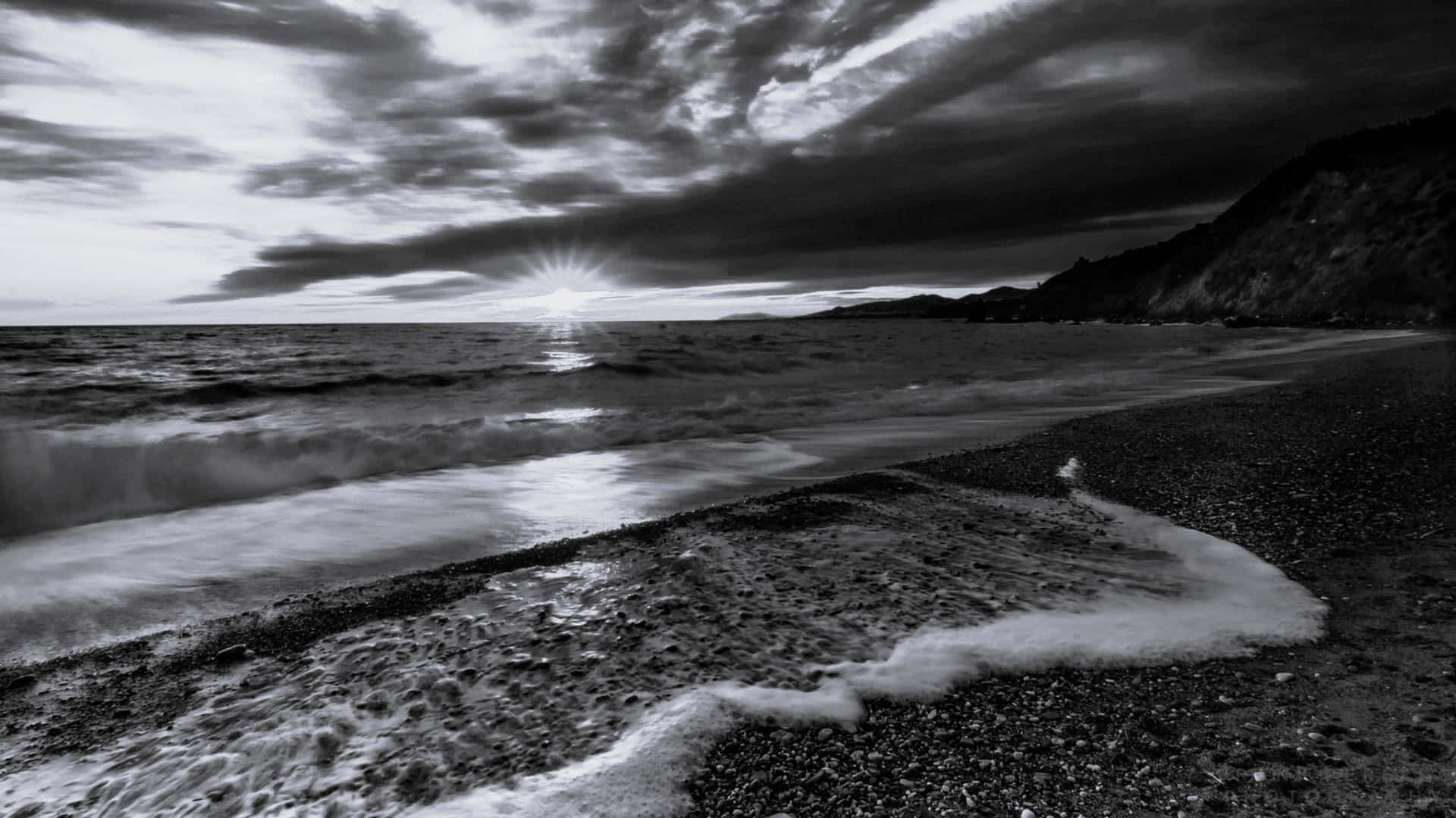  壁紙 — 戲劇性的黑白海岸場景，陽光穿透暴風雲層照射在卵石海灘上。