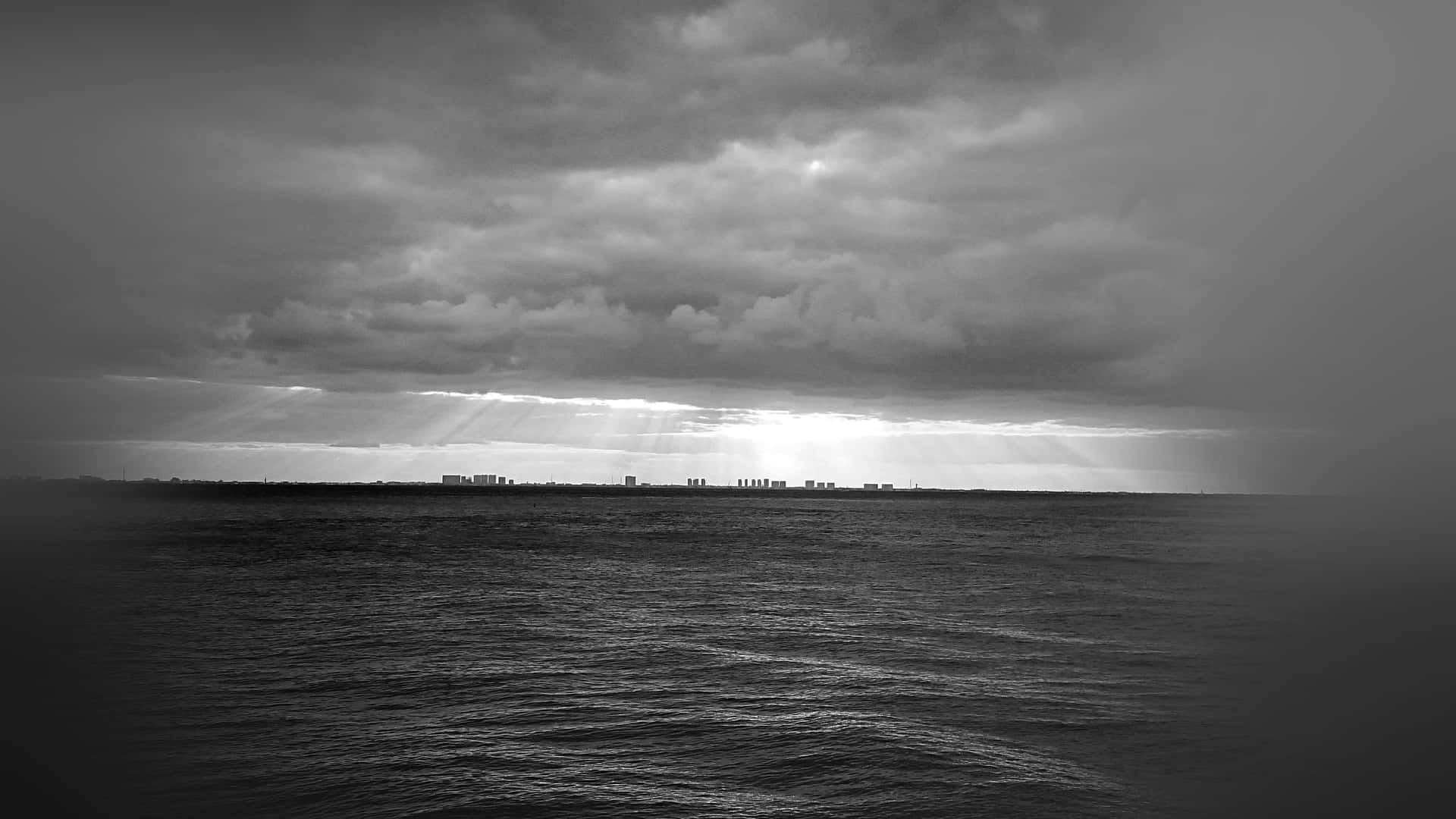  壁紙 — 戲劇性的黑白海景，陽光穿透風暴雲層，照耀在遠處的城市天際線上。