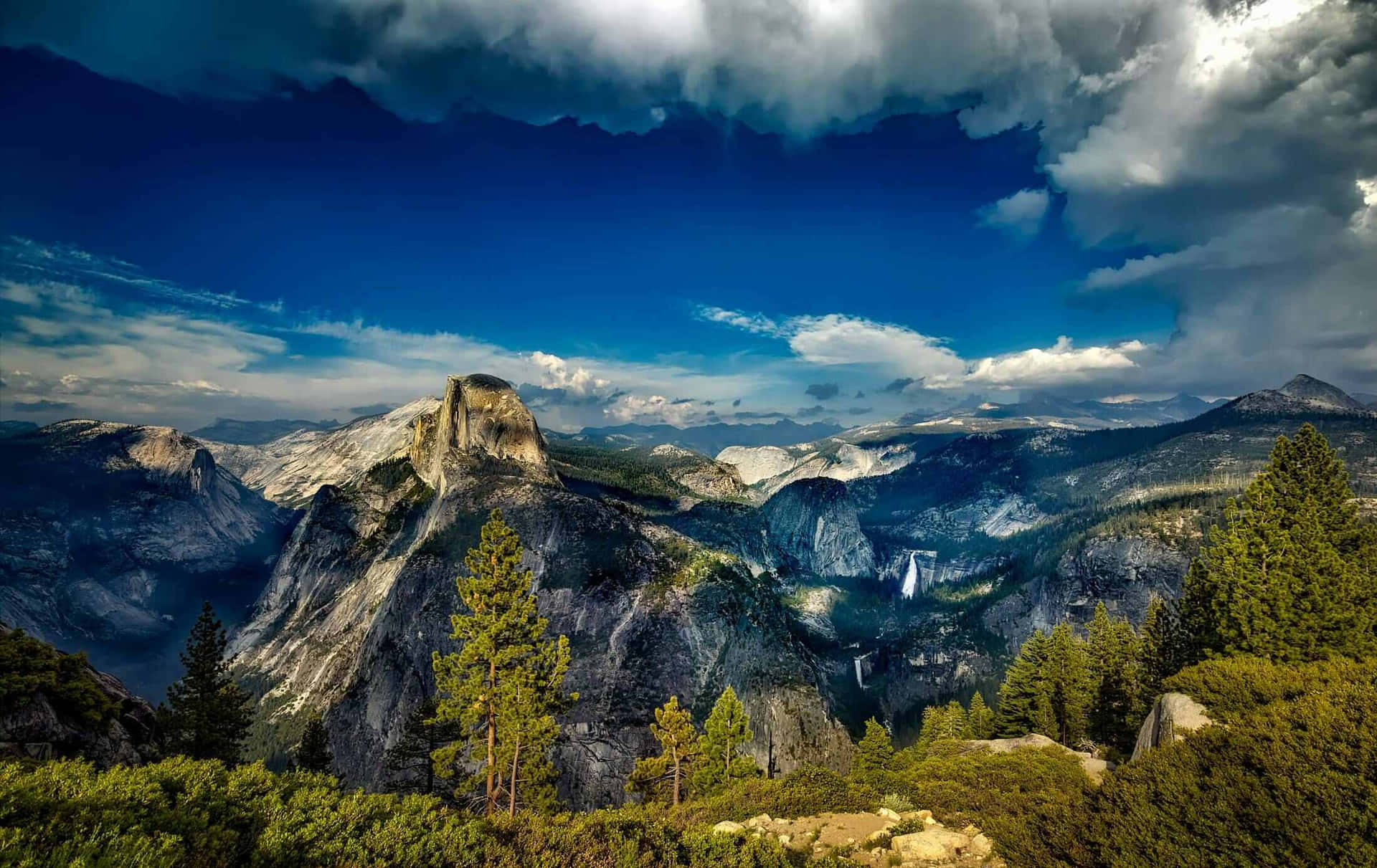  壁紙 — 戲劇性的山景，Half Dome 在風暴般的藍天、散落的雲朵和前景的松樹下聳立。