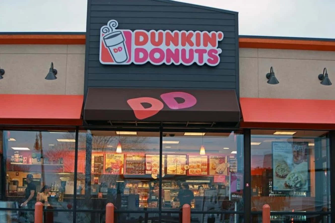 Dunkin Donuts 桌布 壁紙 — Dunkin' Donuts 店鋪外觀，採用大膽的紅色和黑色品牌標誌，大型玻璃窗反射著多雲的天空和室內燈光。